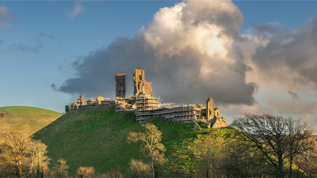 Corfe Castle high in the landscape with scaffolding around the outer walls.  Blue skies with a rain cloud hanging above the castle with a ray of sunshine.
