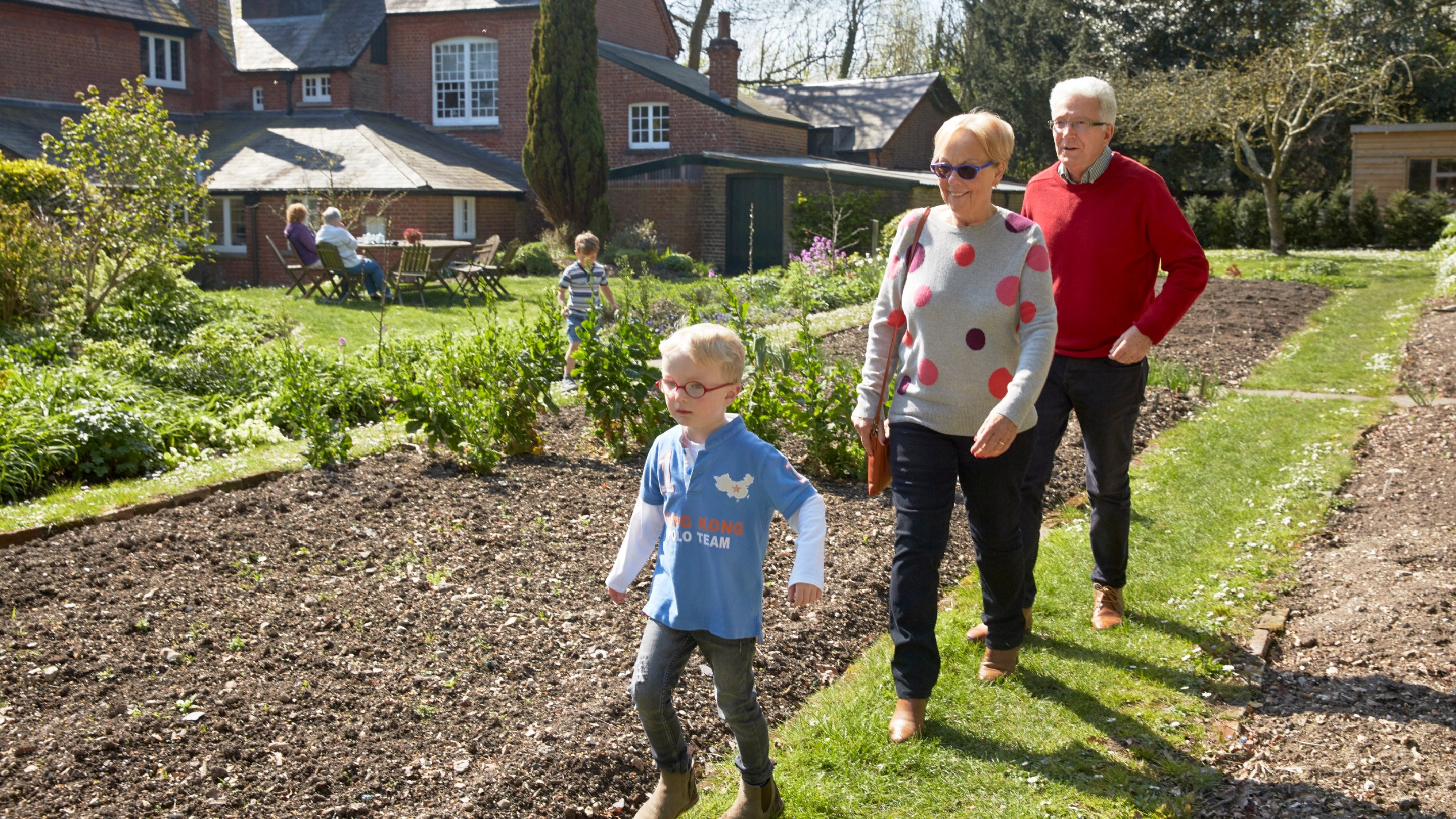 Family members young and old walk on grass paths between flower beds with the red brick house of Max Gate behind