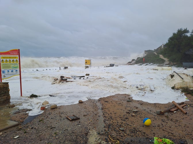 A photograph of Hive Beach during a storm with high waves in the background and debris including a beach ball on the beach