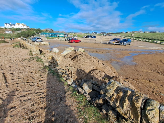 A photograph of Hive Beach car park after a storm, with walls damaged and sand piled up as a result of the storm.