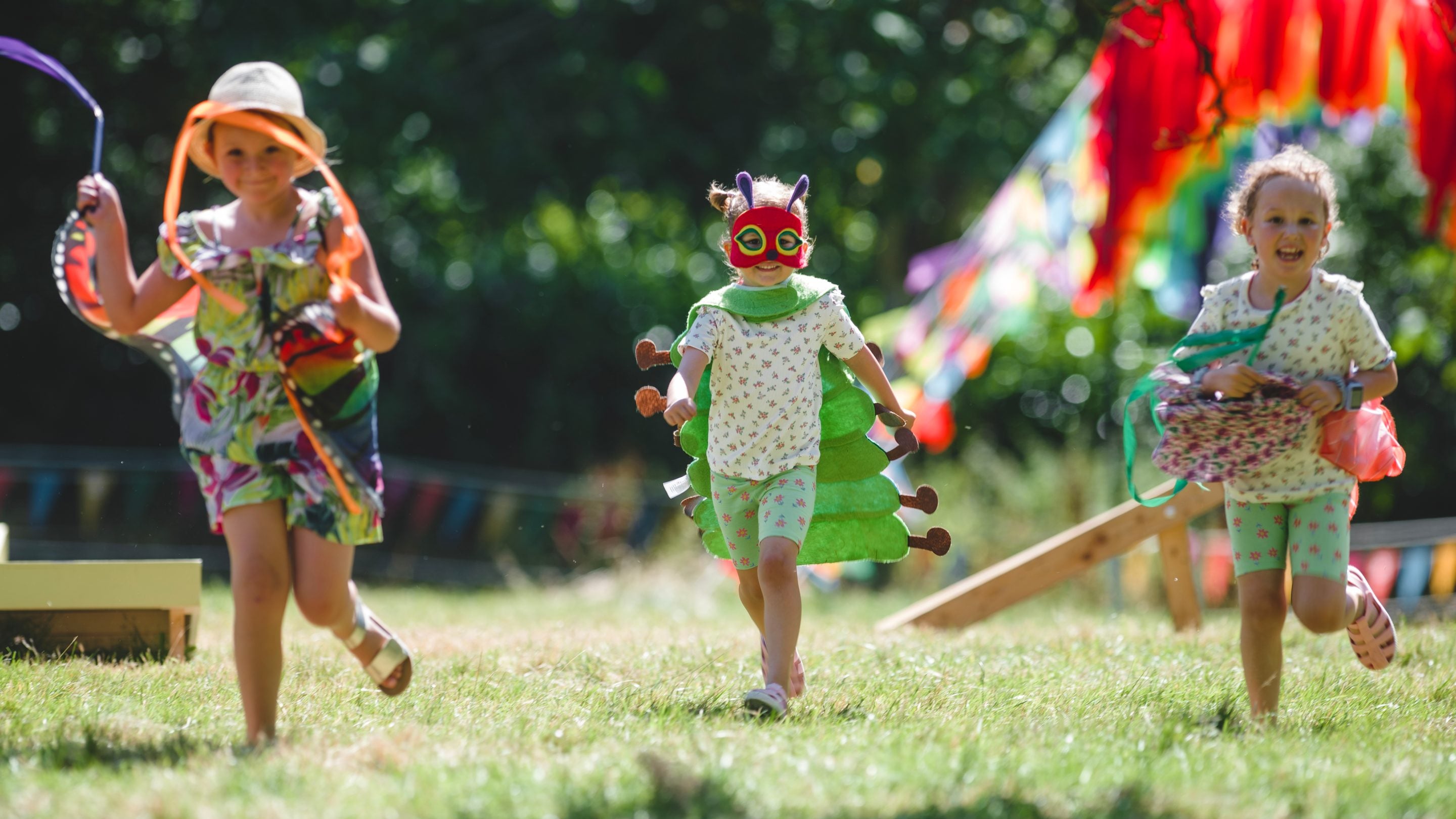 Three children run towards the camera, waving streamers. They are dressed in colourful summer clothes and the sun is shining