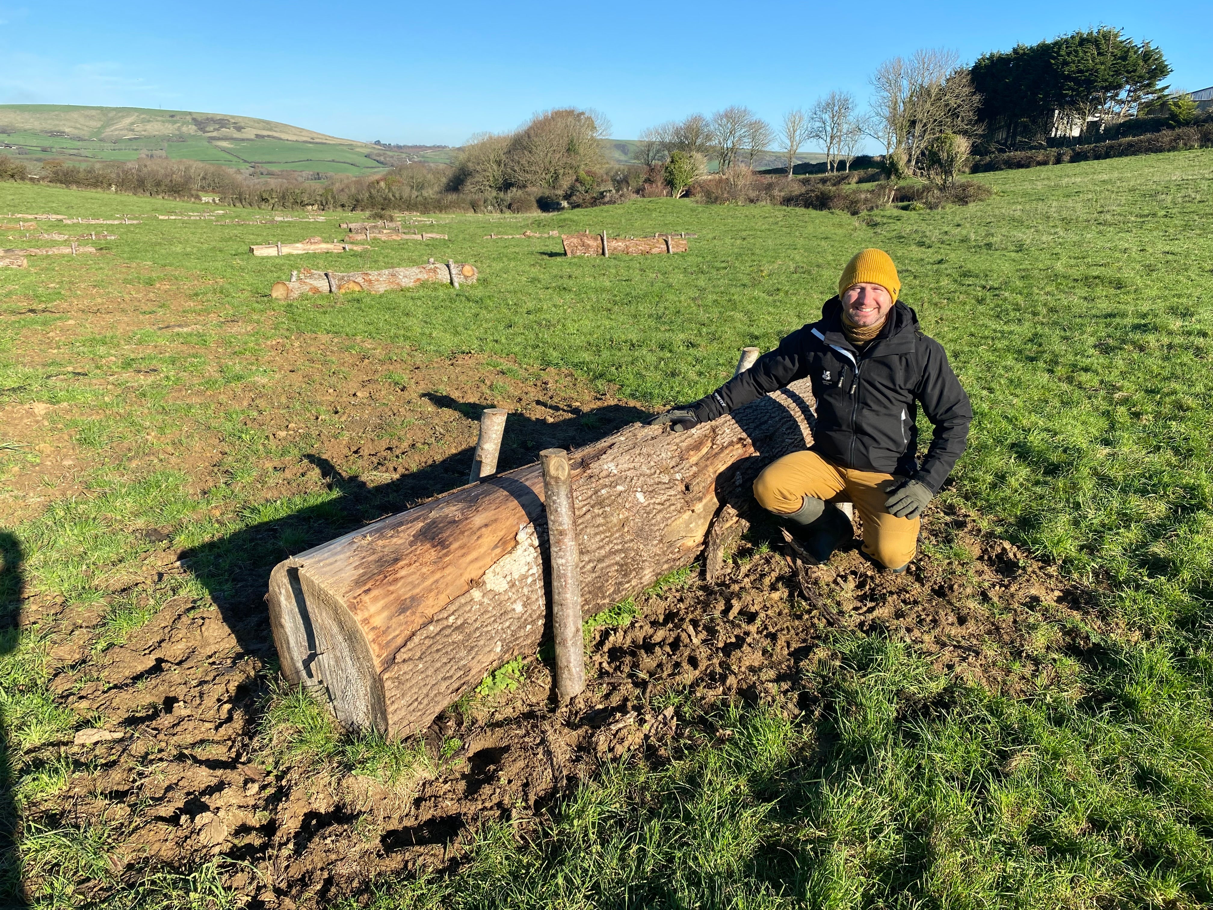 Ranger demonstrating surface roughening - he is crouching beside a log that has been laid in a field to slow down the flow of water. Other logs are laid out behind him.