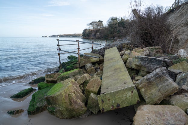 Failing sea defences comprising boulders and broken wire cages full of rocks, with sea lapping against them.
