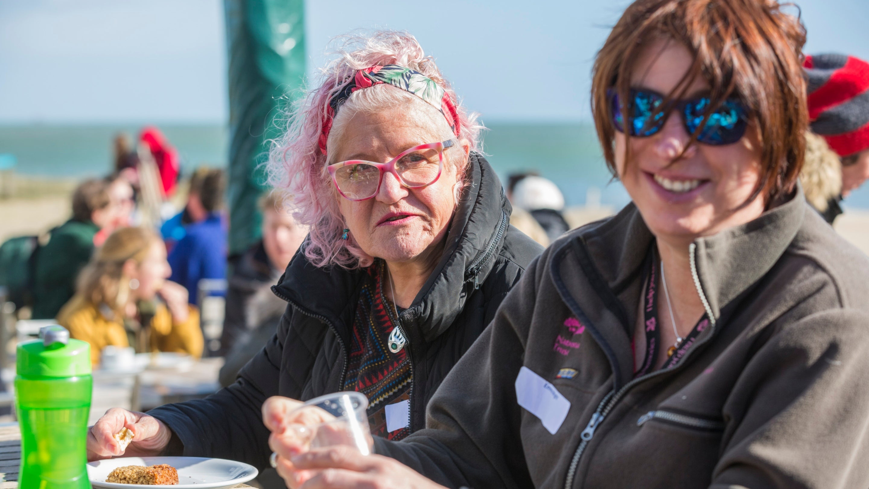 Two volunteers in National Trust jackets sit at a picnic table on Studland Beach, sharing drinks and cake