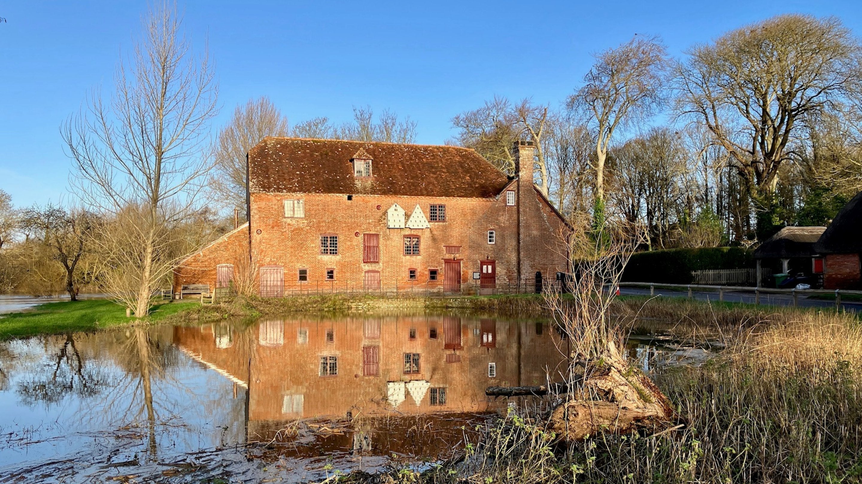 A large brick building in winter sunshine, with a pond in front of it and bare trees behind