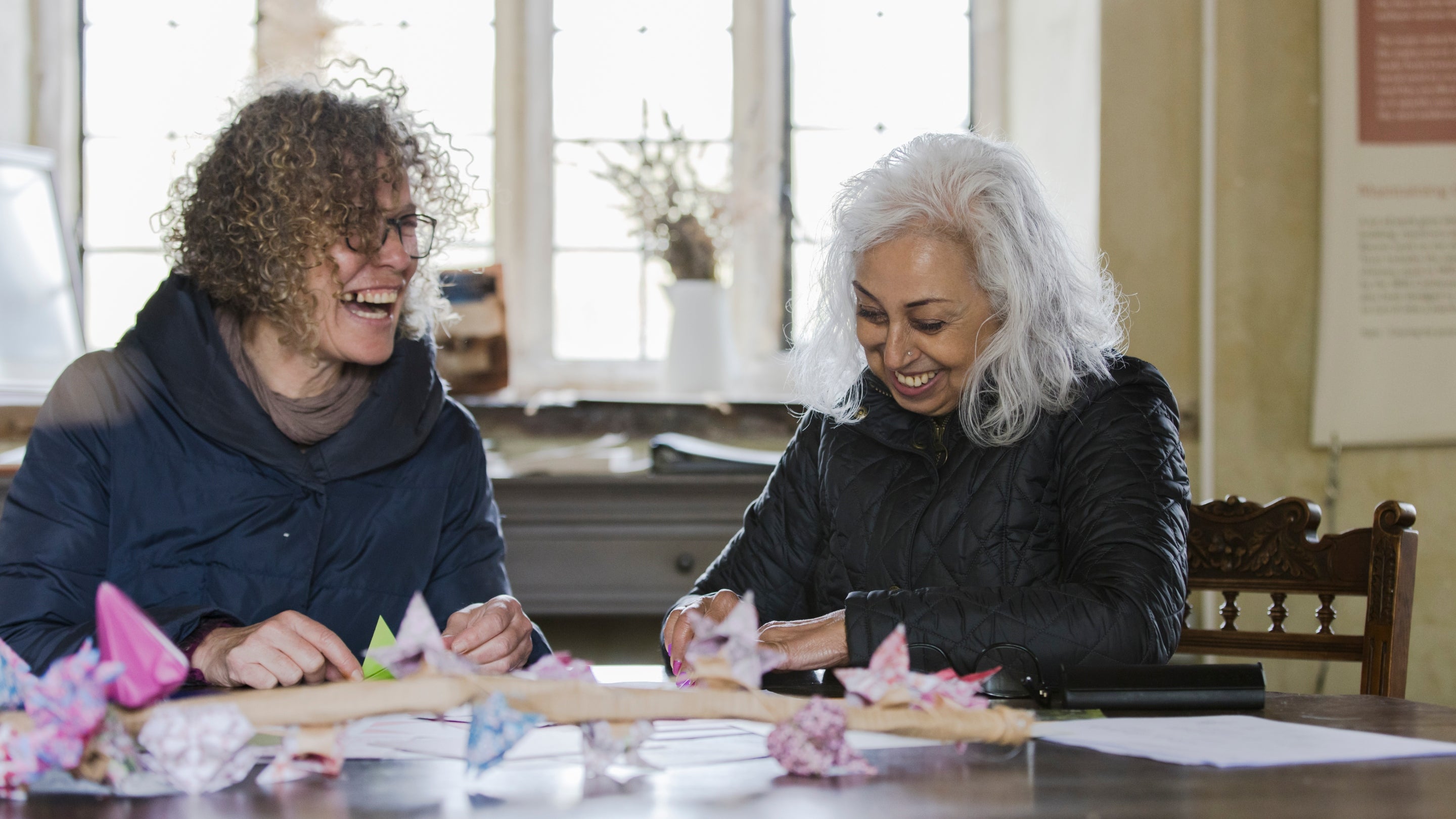 A pair of people sit at a table, talking to each other and laughing while they participate in a blossom origami craft activity.