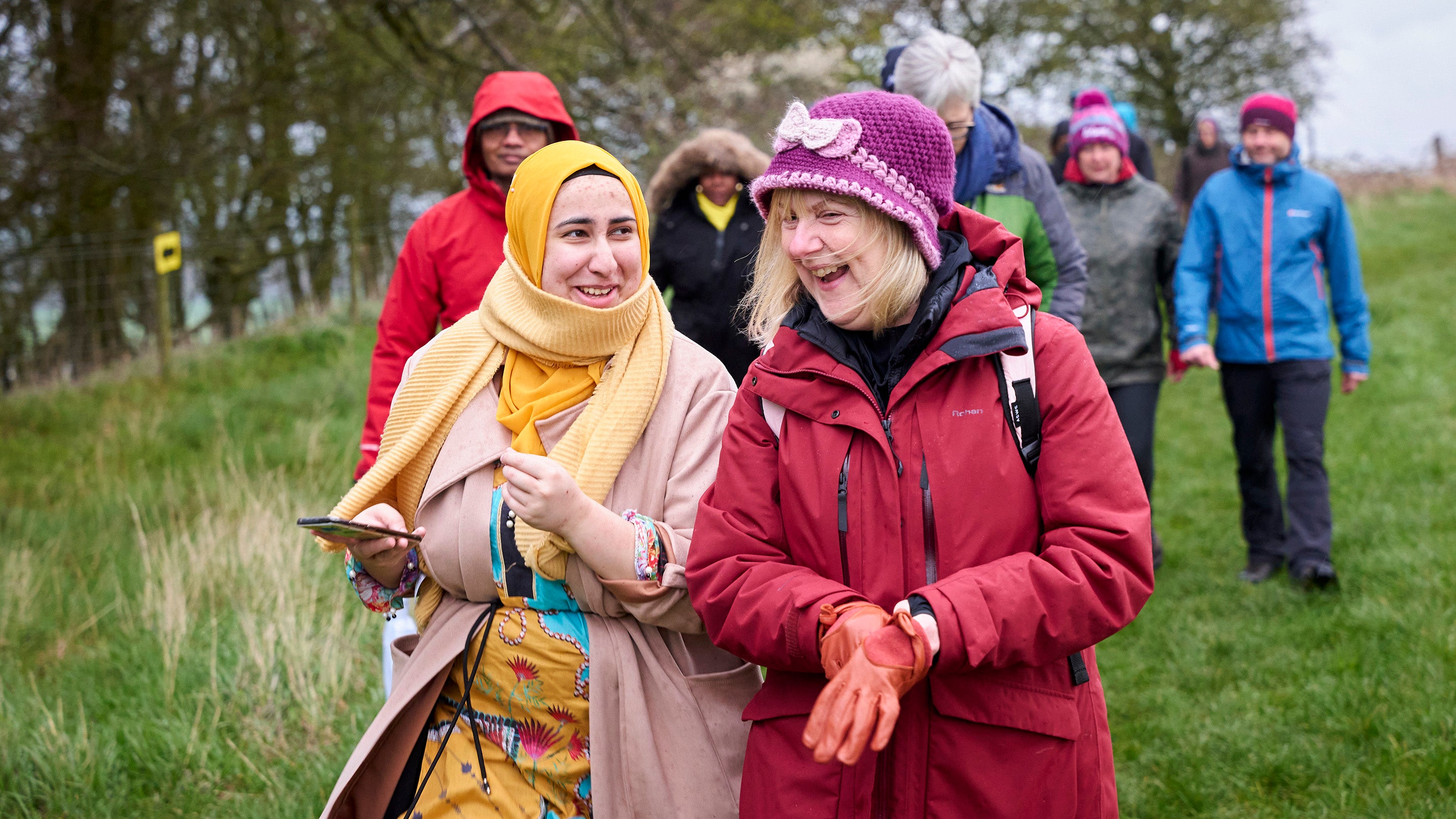 A group of people walking along a field side with a pair of women at the front laughing at each other