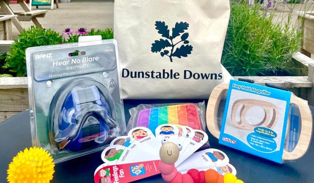 a cotton drawstring bag with the National Trust logo and Dunstable Downs printed on it. Sits on a table surrounded by sensory items: earmuffs, fidget toys, feelings fan and easy hold wooden magnifier.