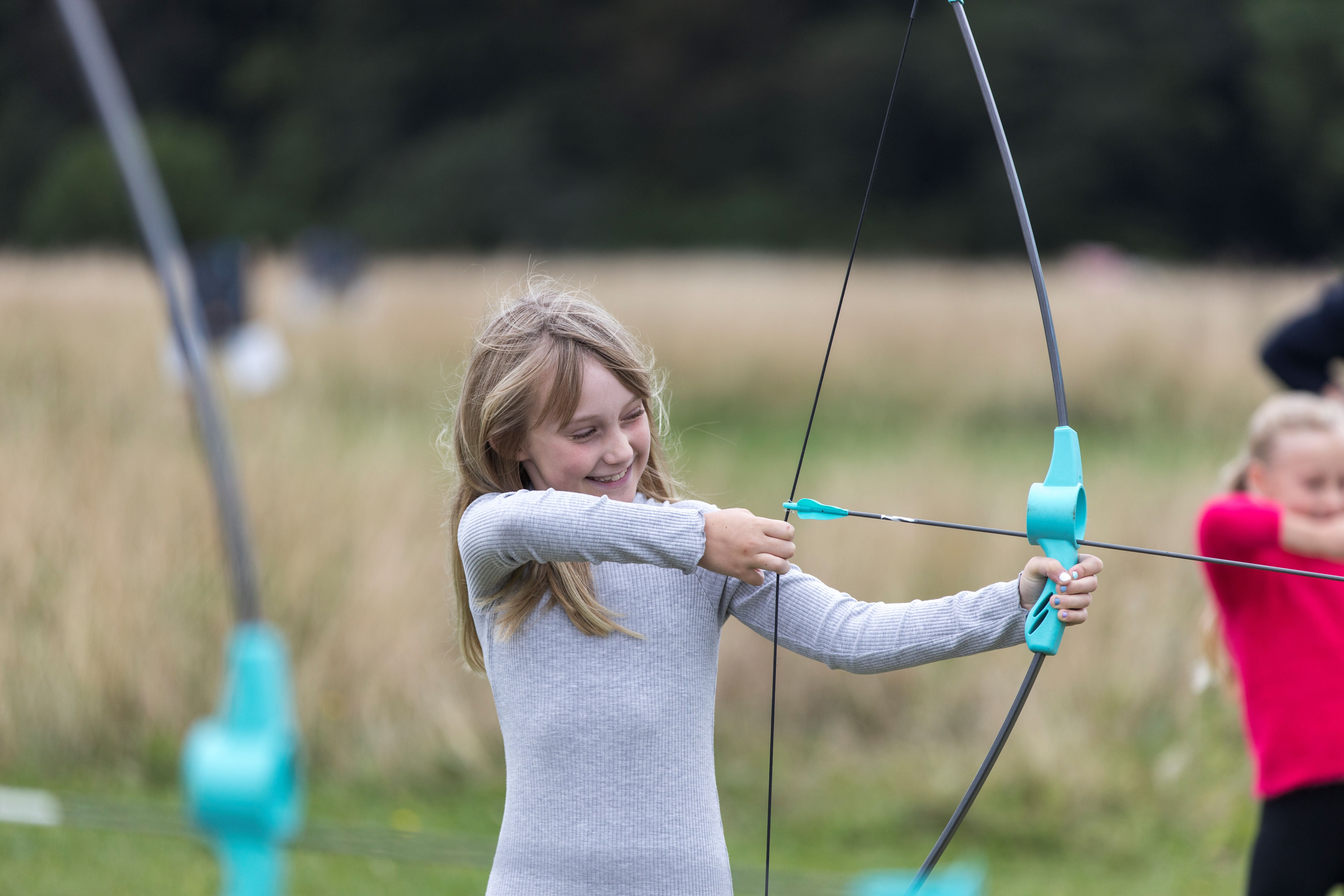 A girl in a grey top smiling while pulling back an archery bow and arrow