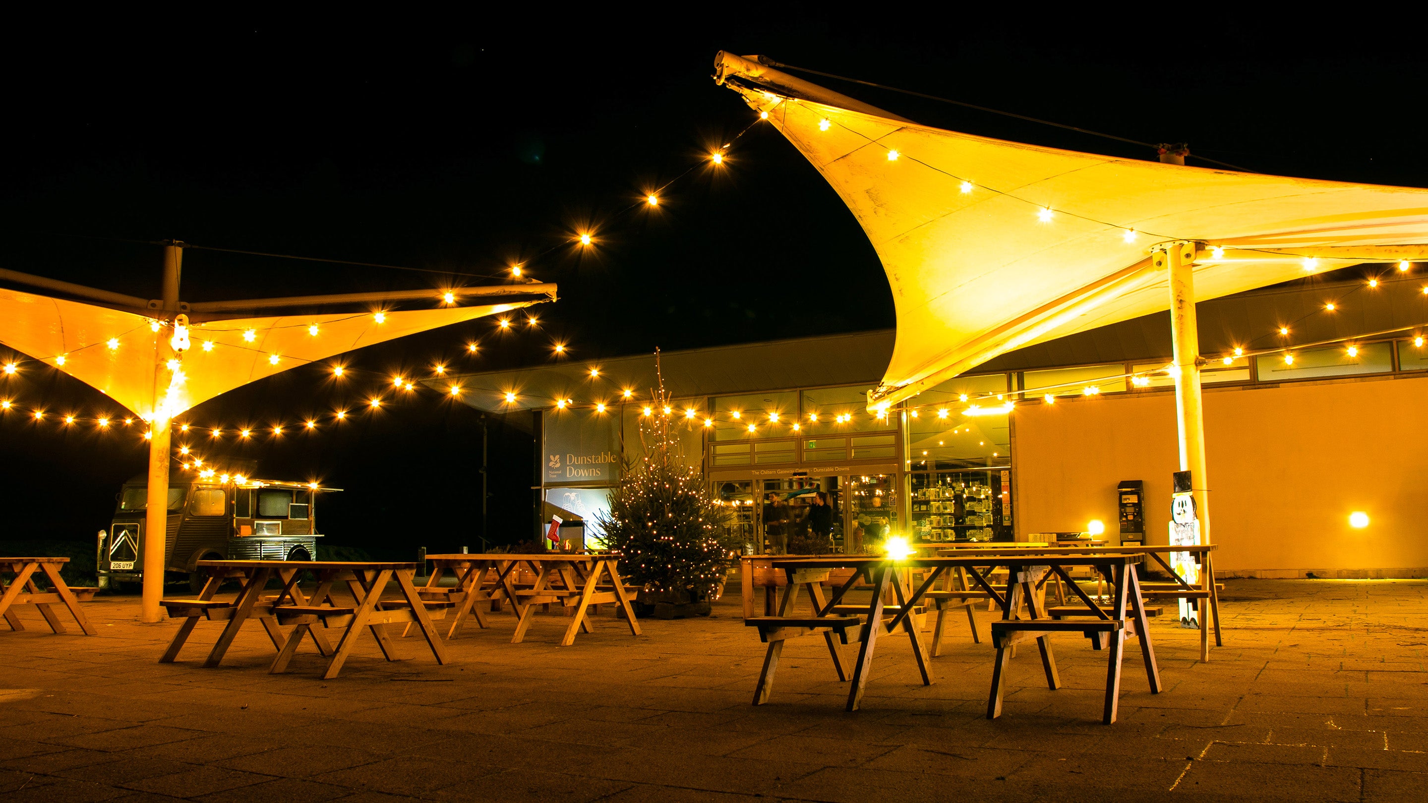 A night time shot of the Dunstable Downs visitor centre with festoon lights strung between two sail covers. There is a lit christmas tree in the centre of the image
