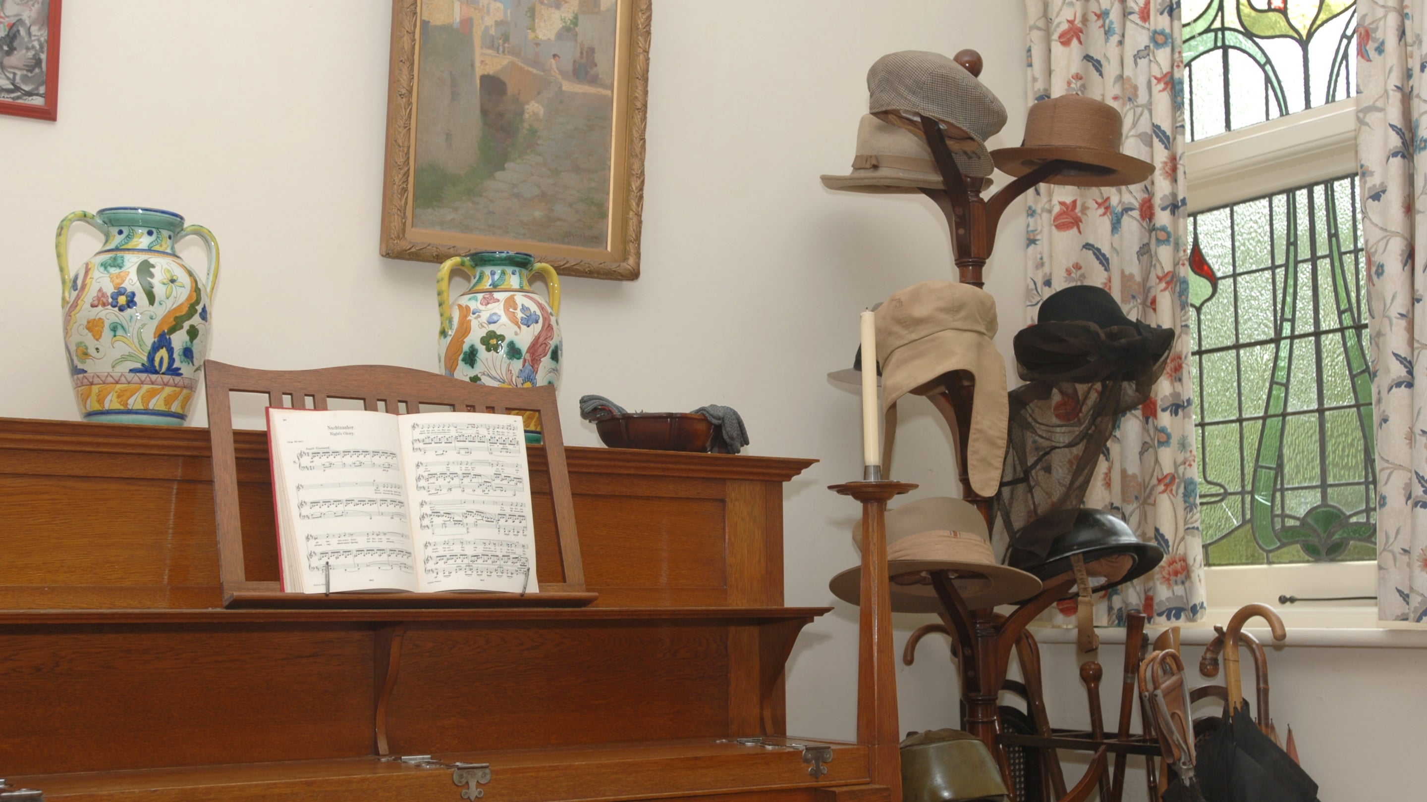 A Bechstein piano and collection of hats in the Entrance Hall at Shaw's Corner, Hertfordshire. Sheet music is visible on the piano and the hats are hung on an upright hat stand.