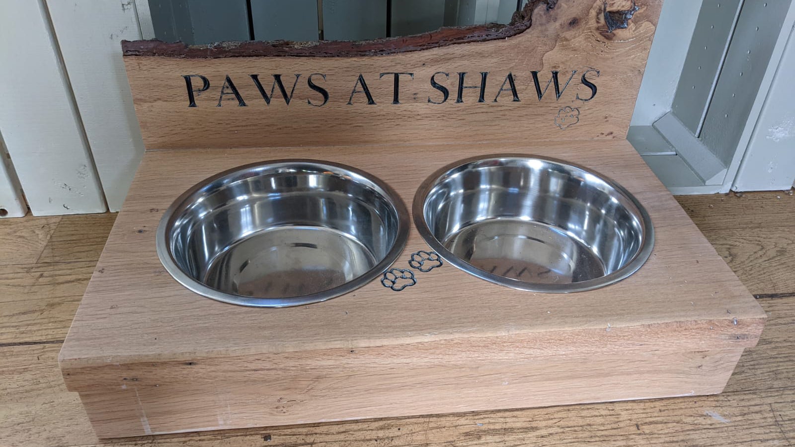 a wooden stand with two stainless steel dog bowls in it. On the back Shaws Paws is carved into the wood