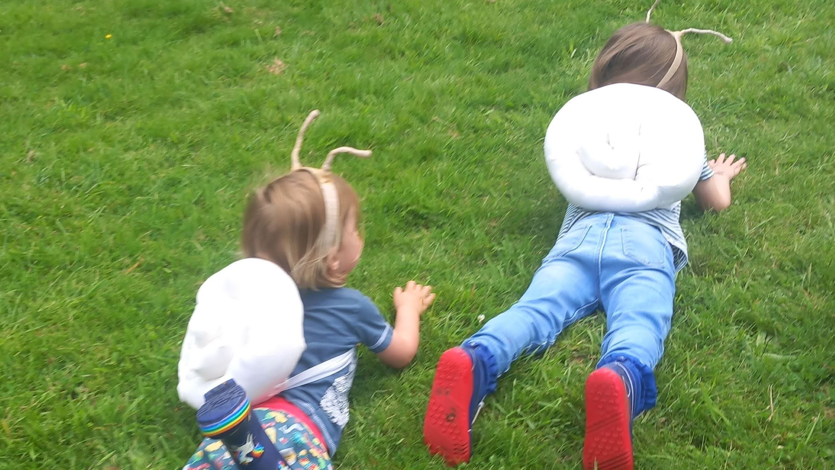 Two children with "snail shells" strapped to their backs and antennae crawling on their bellies on the grass pretending to be snails