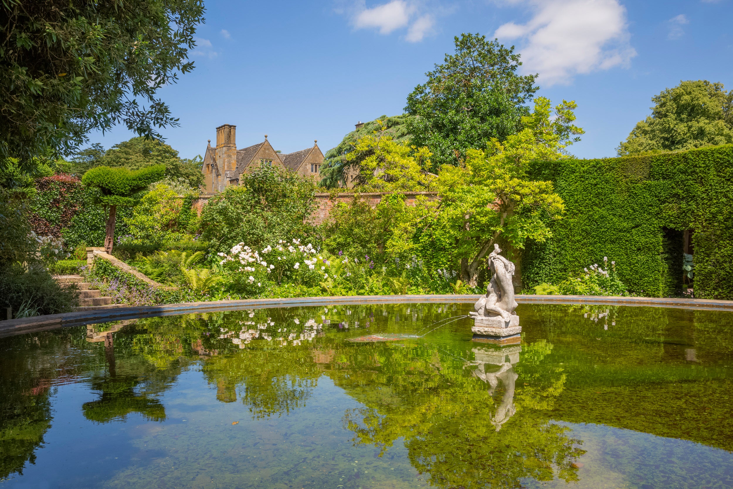 A large circular pond with a stone fountain in the centre and a garden with trees and a manicured hedge behind, an old manor house in the background.