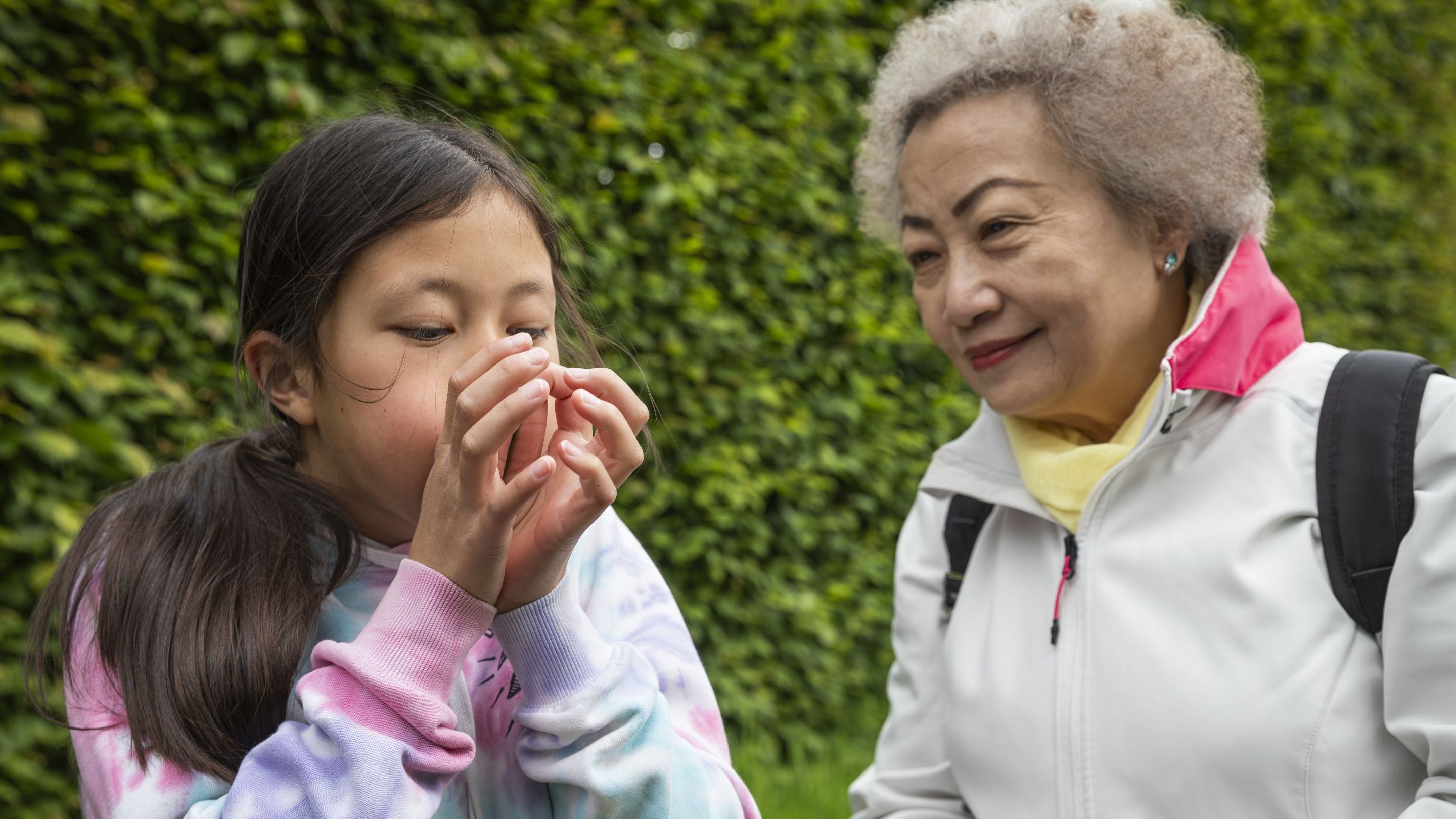 Child blows into a blade of grass in their hands while an older adult looks on, standing outdoors in front of green foliage.