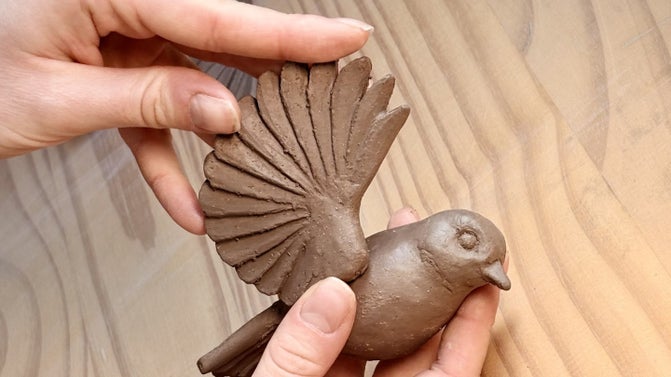 Hands attaching a ceramic wing to a sculpture of a clay bird