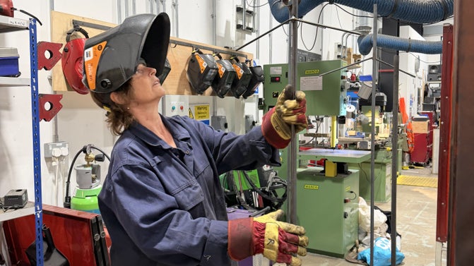 Woman in blue overalls and welder's mask looking upward in a semi-industrial setting
