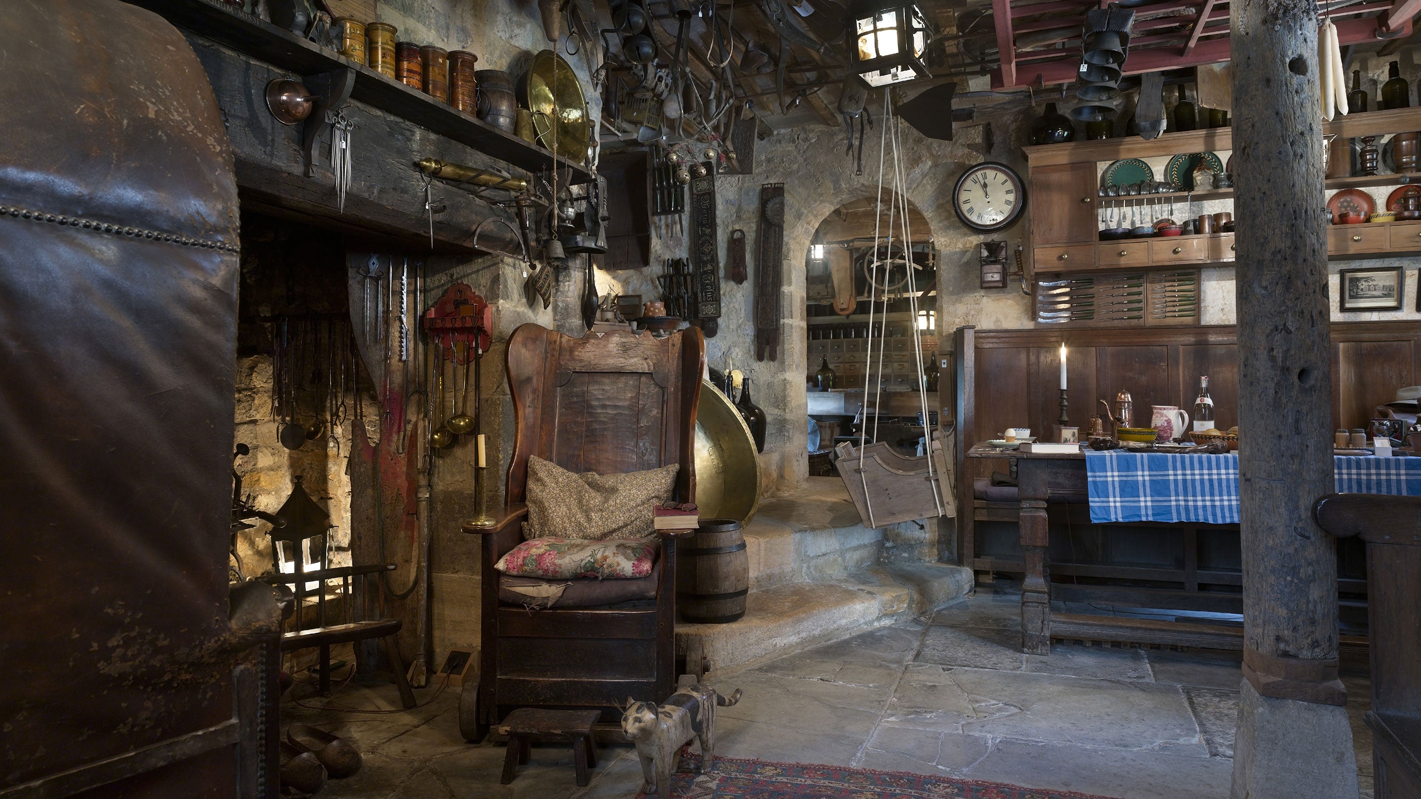 Interior view of a cluttered, rustic living space. An old wooden wingback chair sits in front of a large fireplace, a refectory table is to the left of the chair, using old church pews and a swing as seating.