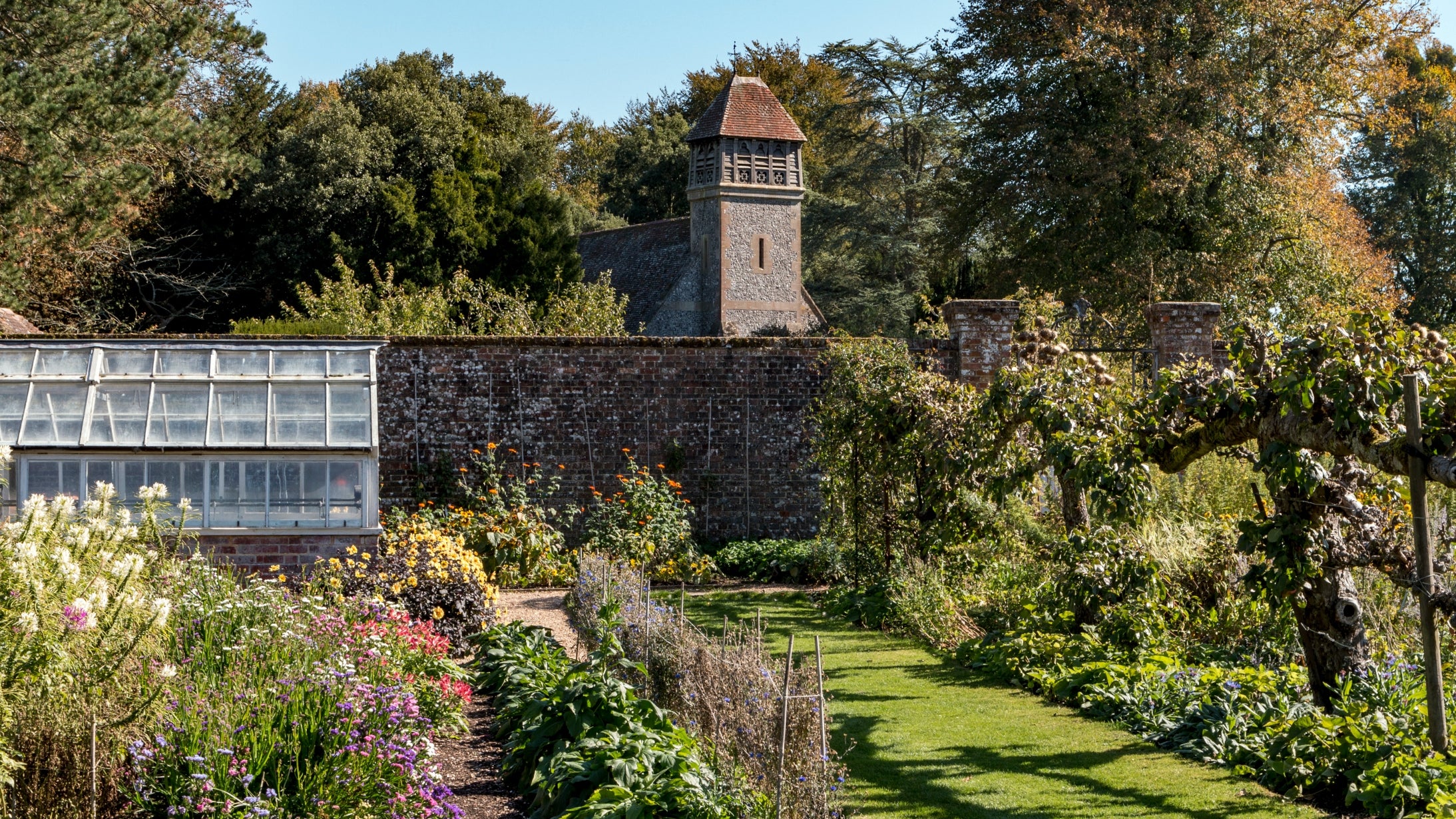 Walled kitchen garden with flower borders, glasshouse, grass paths, and church behind wall in background