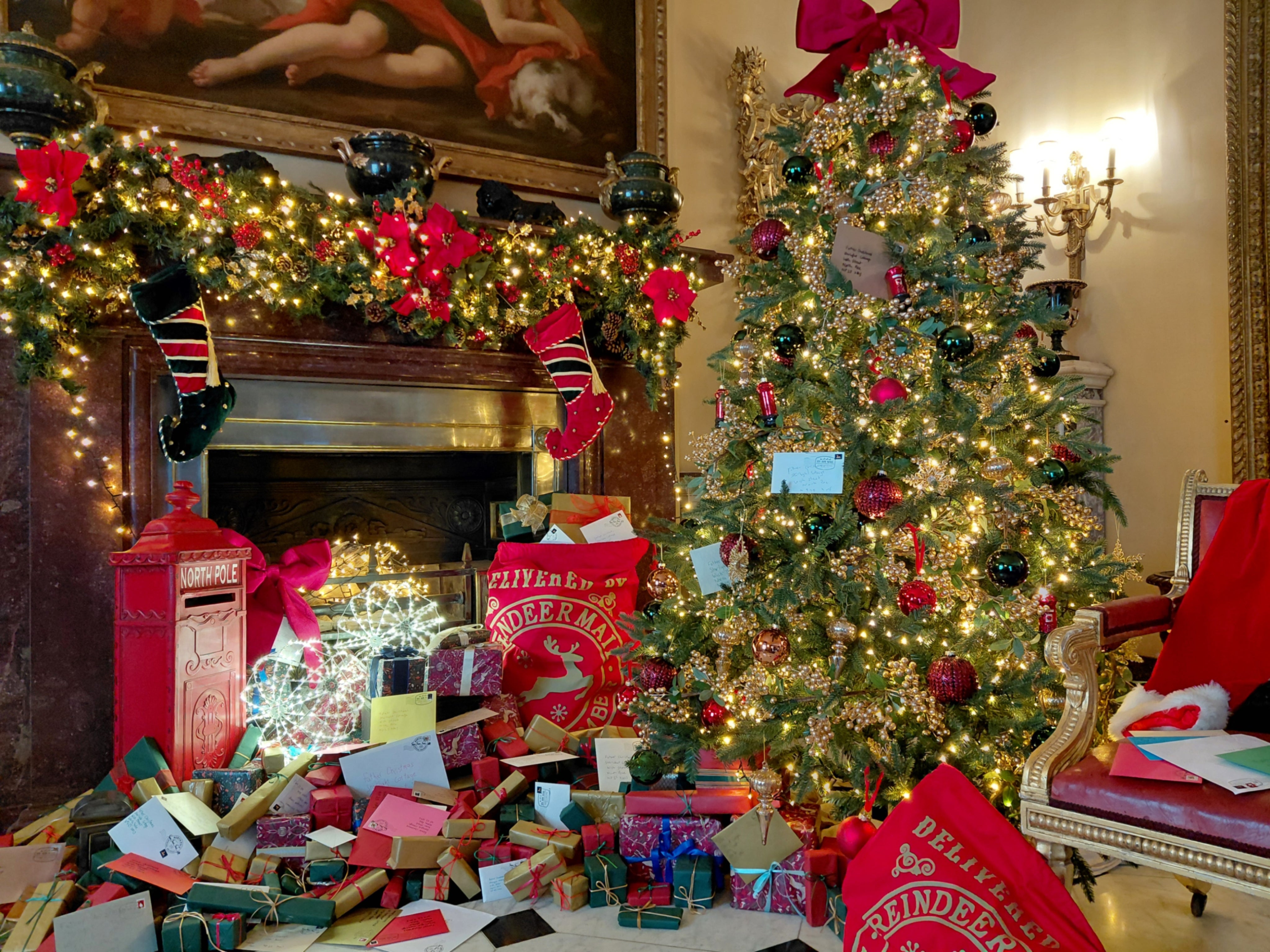 Festively decorated room featuring a large Christmas tree adorned with twinkling lights, red and gold ornaments, and letters to Father Christmas, beside a garland-draped fireplace with stockings, a North Pole post box, sacks of Christmas mail, and piles of wrapped presents.