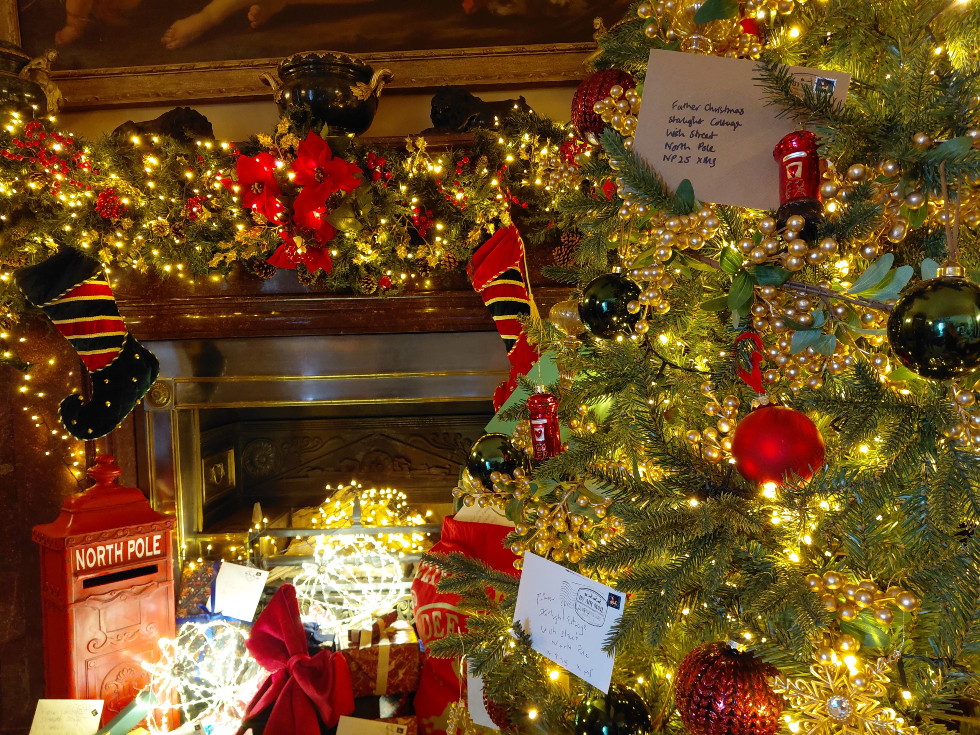 Decorated Christmas tree beside a fireplace, illuminated with warm white lights and adorned with red and black baubles, gold berry garlands, stockings, wrapped presents, and letters to Father Christmas, creating a festive holiday display.