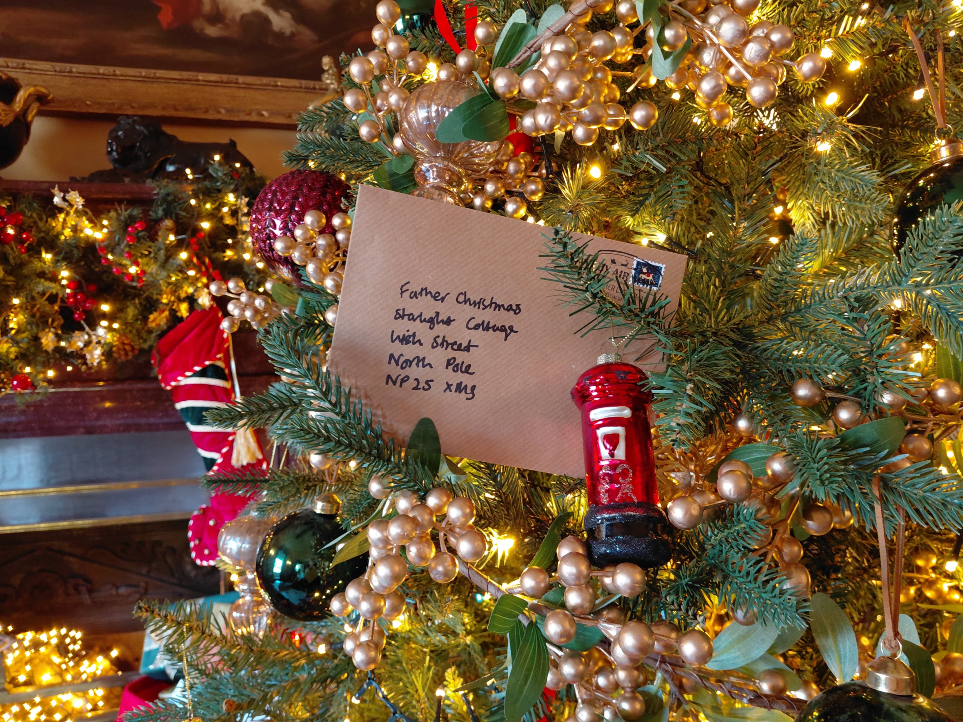 A Christmas tree decorated with warm white lights, gold berry garlands and colourful baubles, featuring a handwritten letter addressed to Father Christmas nestled among the branches, with a festive fireplace and Christmas stocking in the background.