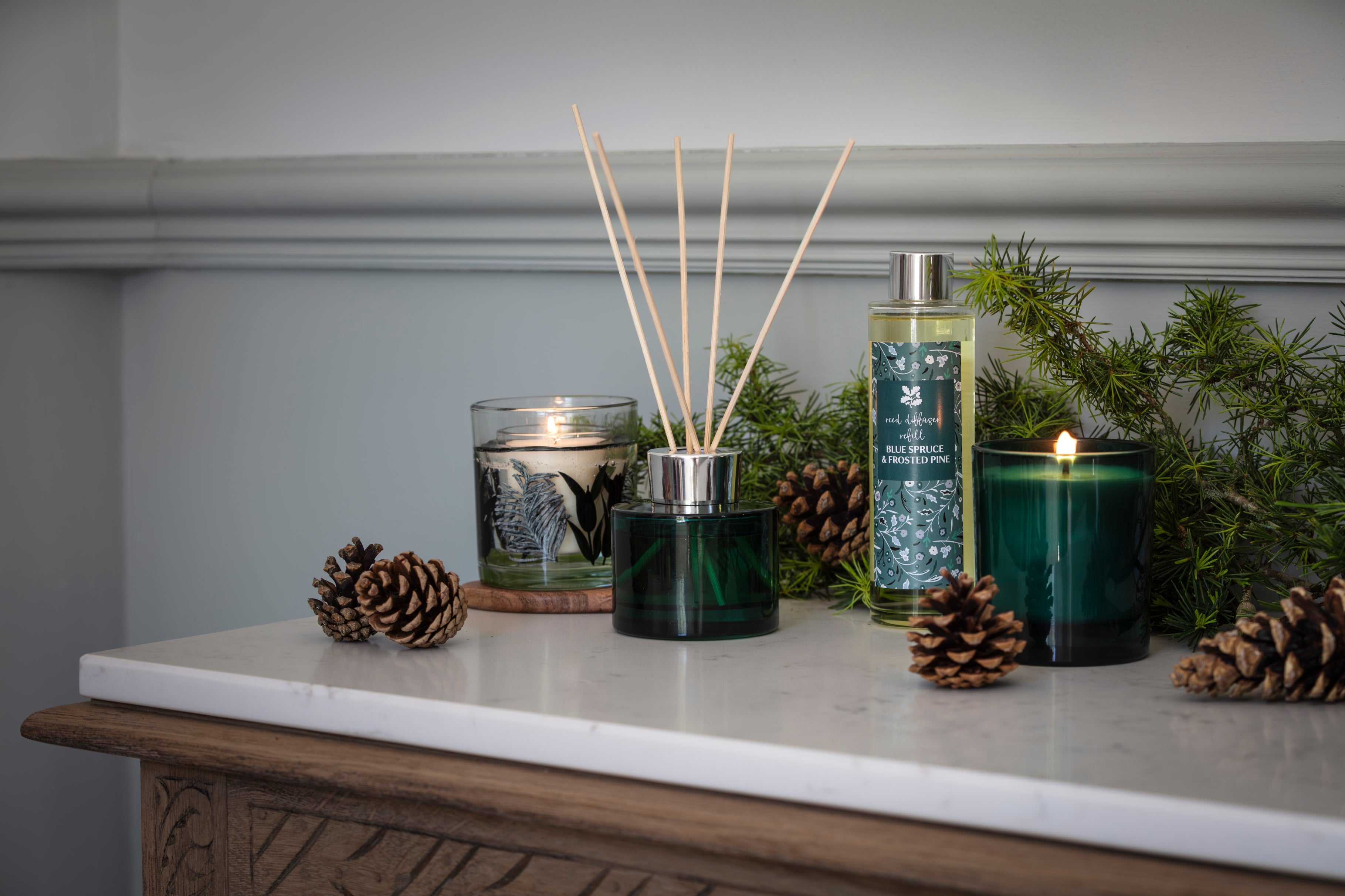 Autumn home fragrance collection with candles, reed diffuser, pine cones and evergreen foliage on a marble-topped table.