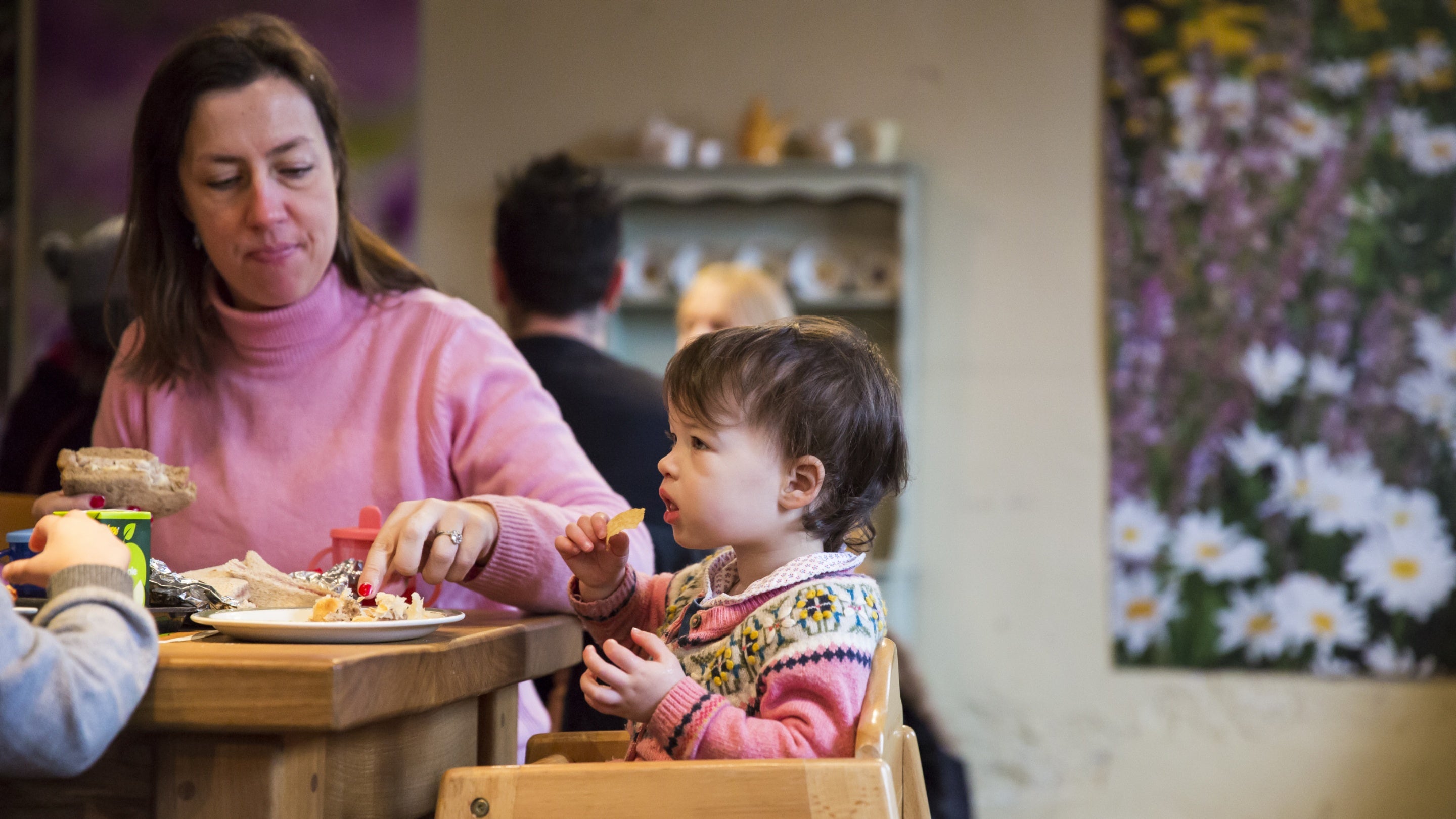 A woman in a pink jumper, eating a sandwich, feeds a toddler in a chunky knit cardigan, sitting in a wooden high chair in a café