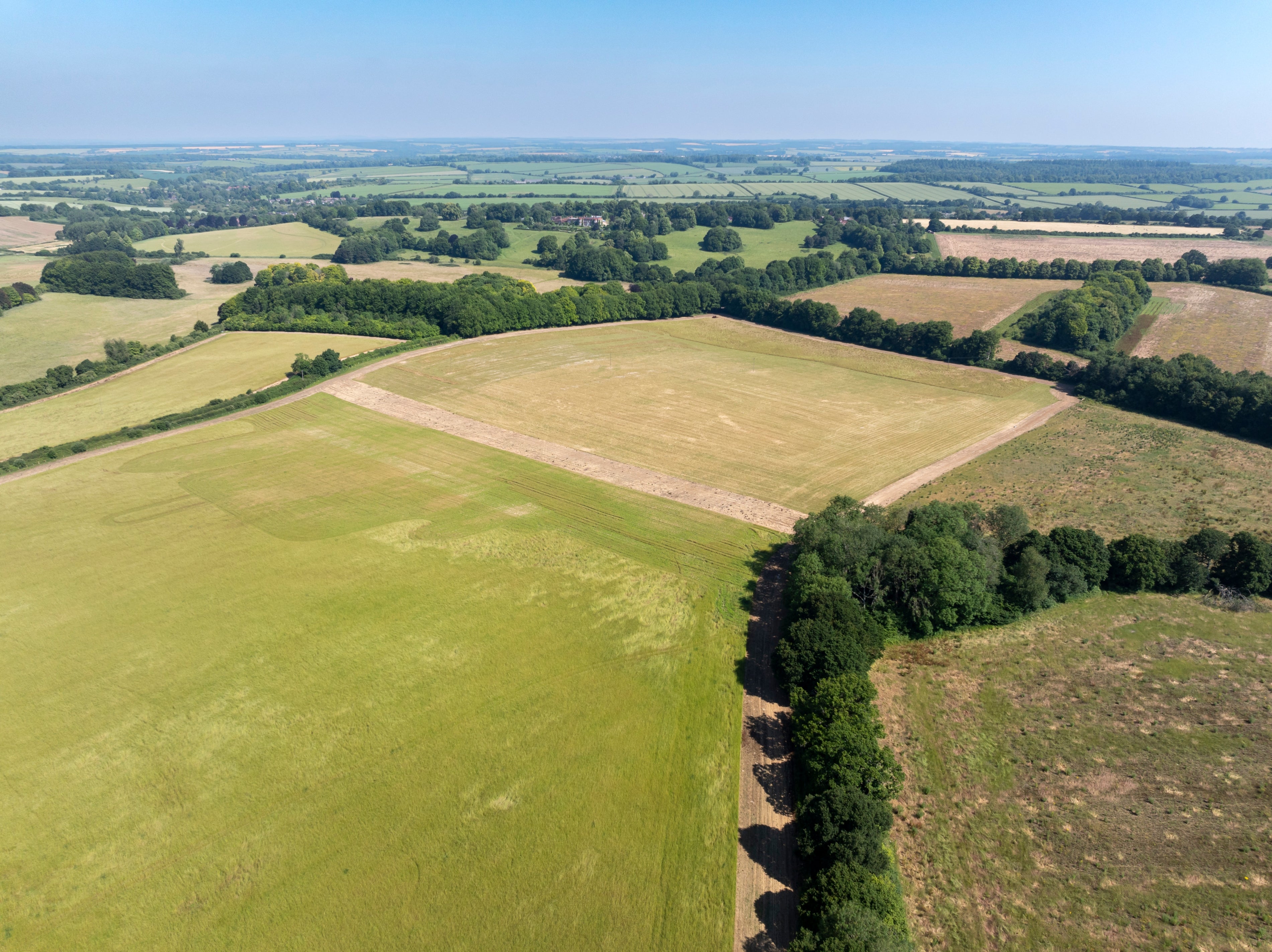 A high-angle drone photograph showing a patchwork of agricultural fields and open countryside. Large rectangular fields in shades of green and golden brown are bordered by hedgerows and clusters of mature trees. A dirt track curves around the edge of a harvested field in the foreground, while rolling farmland stretches to the horizon beneath a clear blue sky.