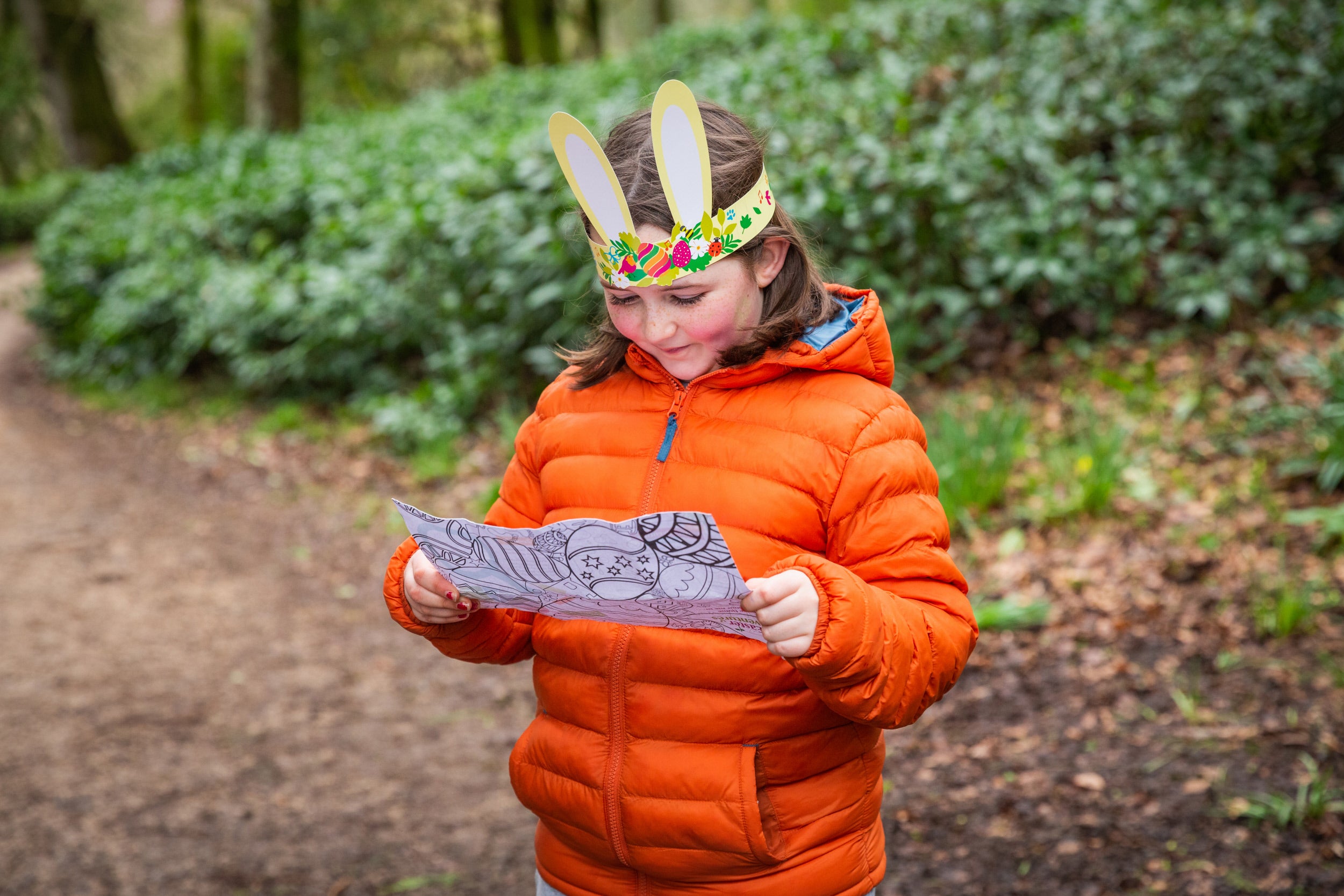 A girl reading her Easter trail activity sheet, wearing yellow bunny ears and an orange jacket