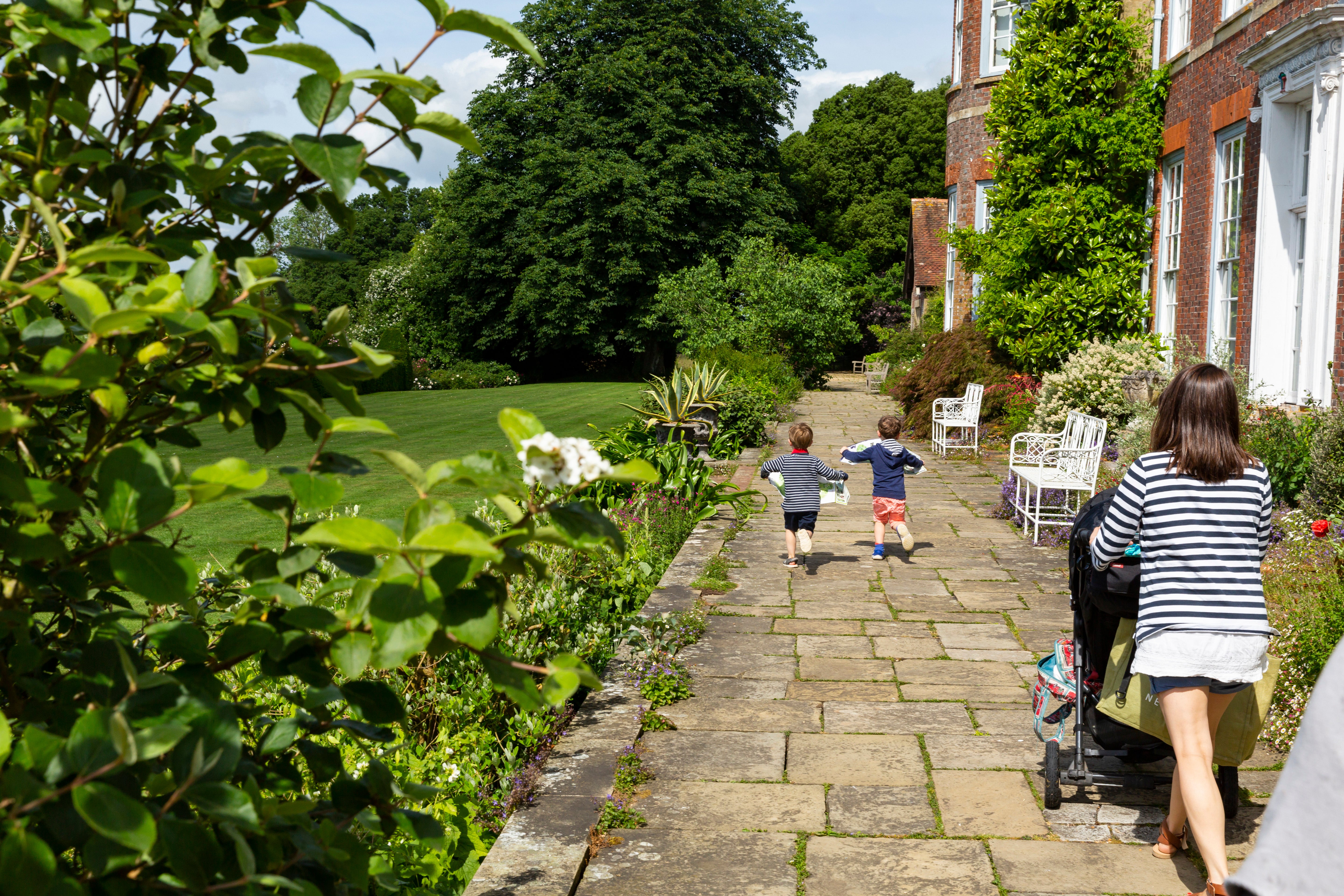A paved garden path runs alongside a brick house with white doors and windows, bordered by lush plants and flowers. Two children run ahead along the path while an adult pushes a stroller behind them, with a large green lawn and tall trees stretching out to the left in bright summer sunshine.