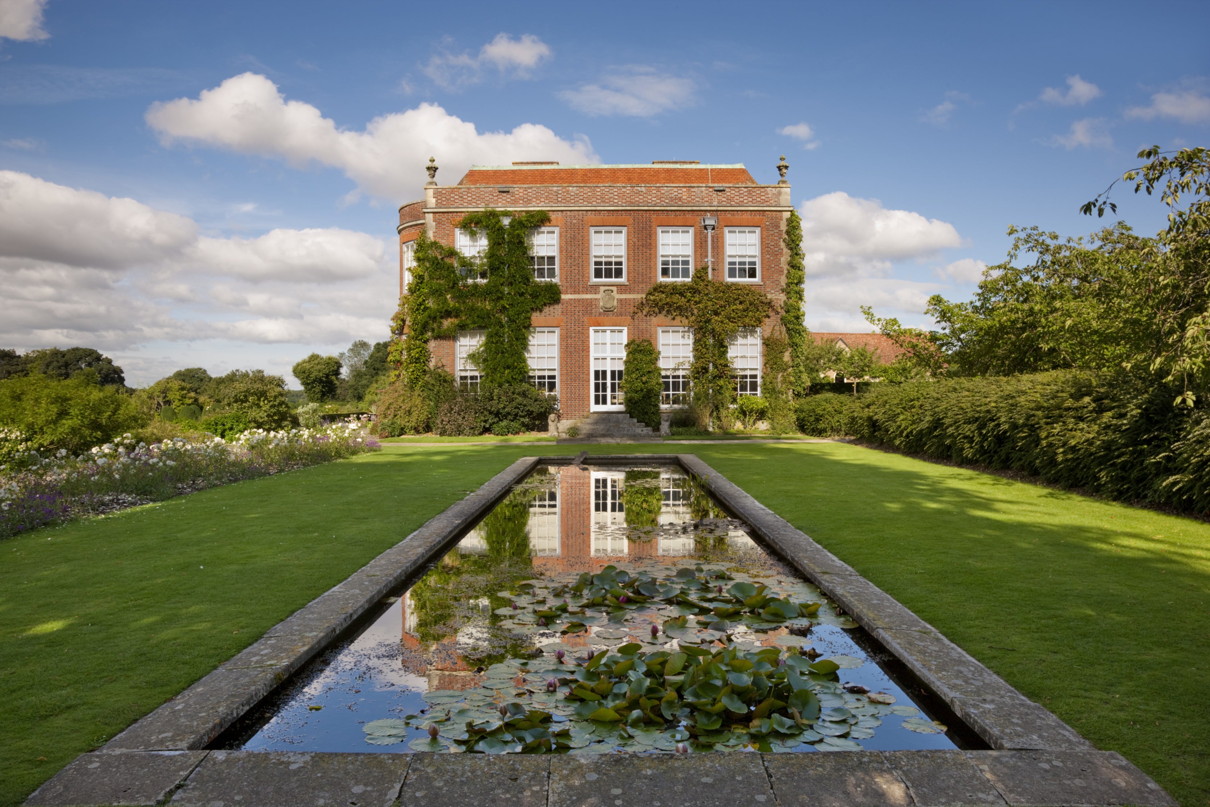Georgian-style red brick house covered in climbing ivy, viewed from a formal garden with a long rectangular reflecting pool filled with lily pads in the foreground, mirrored in the still water, under a bright blue sky with scattered clouds.