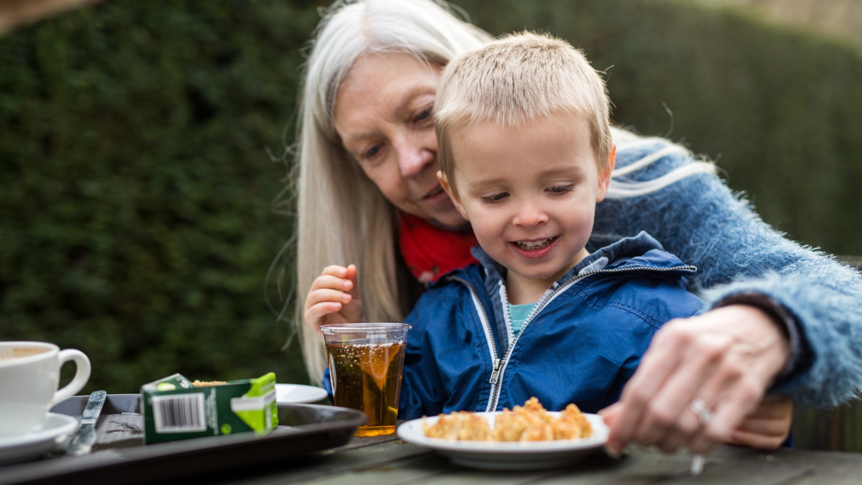 A grandparent and child wearing warm coats sit at a wooden picnic table outside, eating from a plate