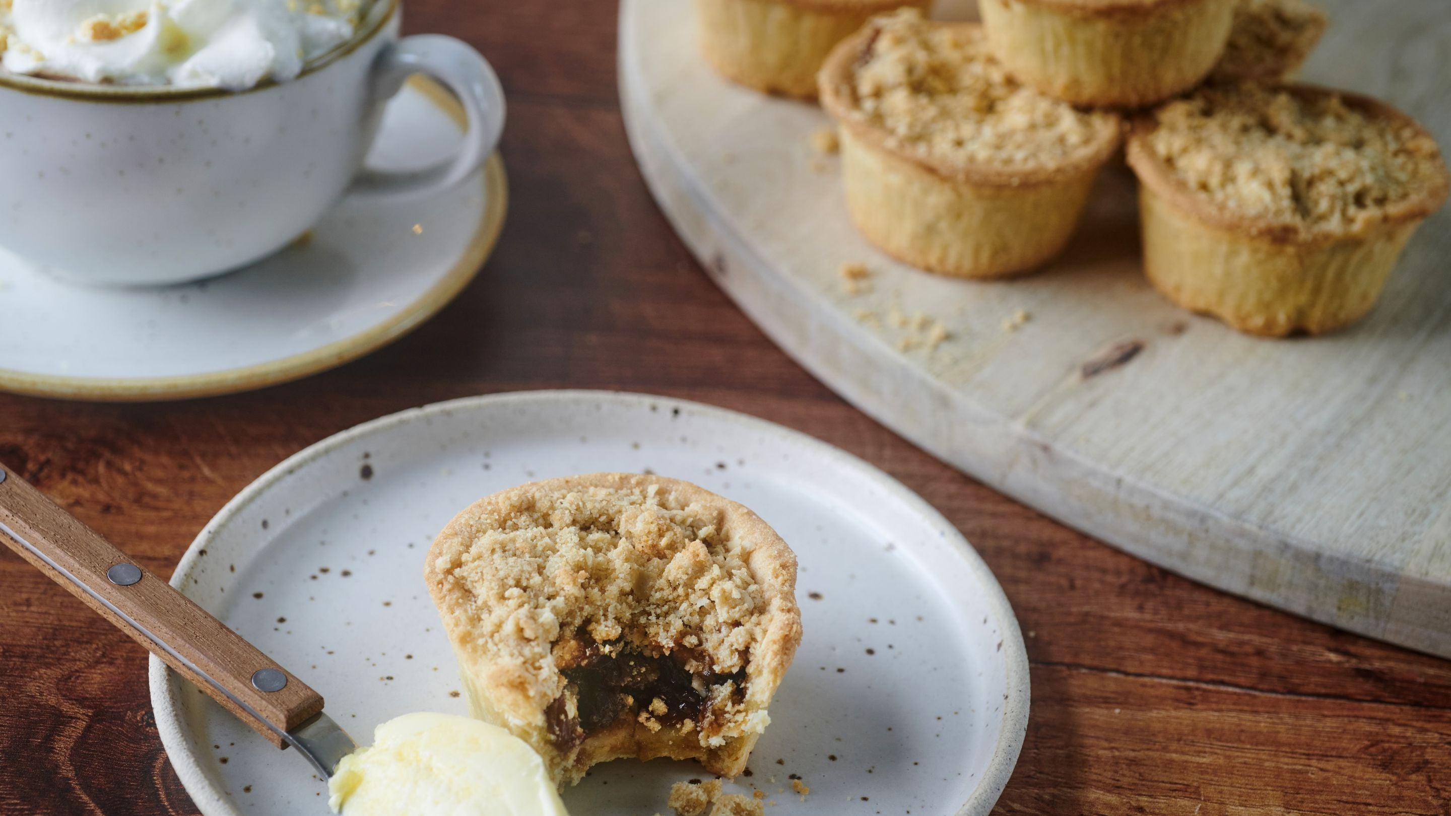 Crumble topped mince pie with a spoonful of clotted cream (or brandy butter) with a cup and saucer on the LHS and a tray of untouched mince pied on the right.