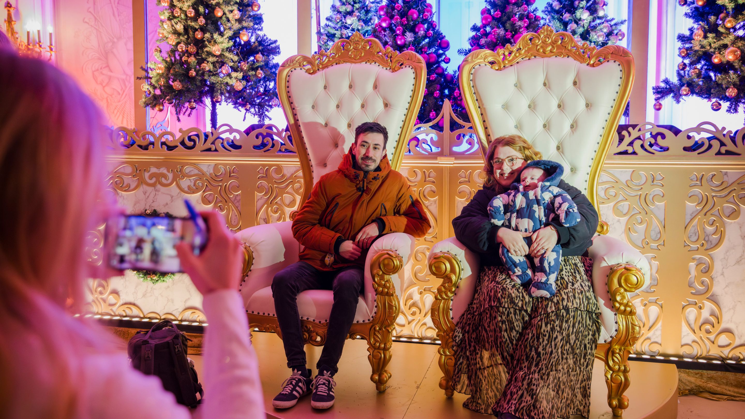 A man with a baby and a woman sit on two large cream and gold thrones, with a raised stage of decorated Christmas trees behind them, for Mottisfont's A Cinderella Christmas