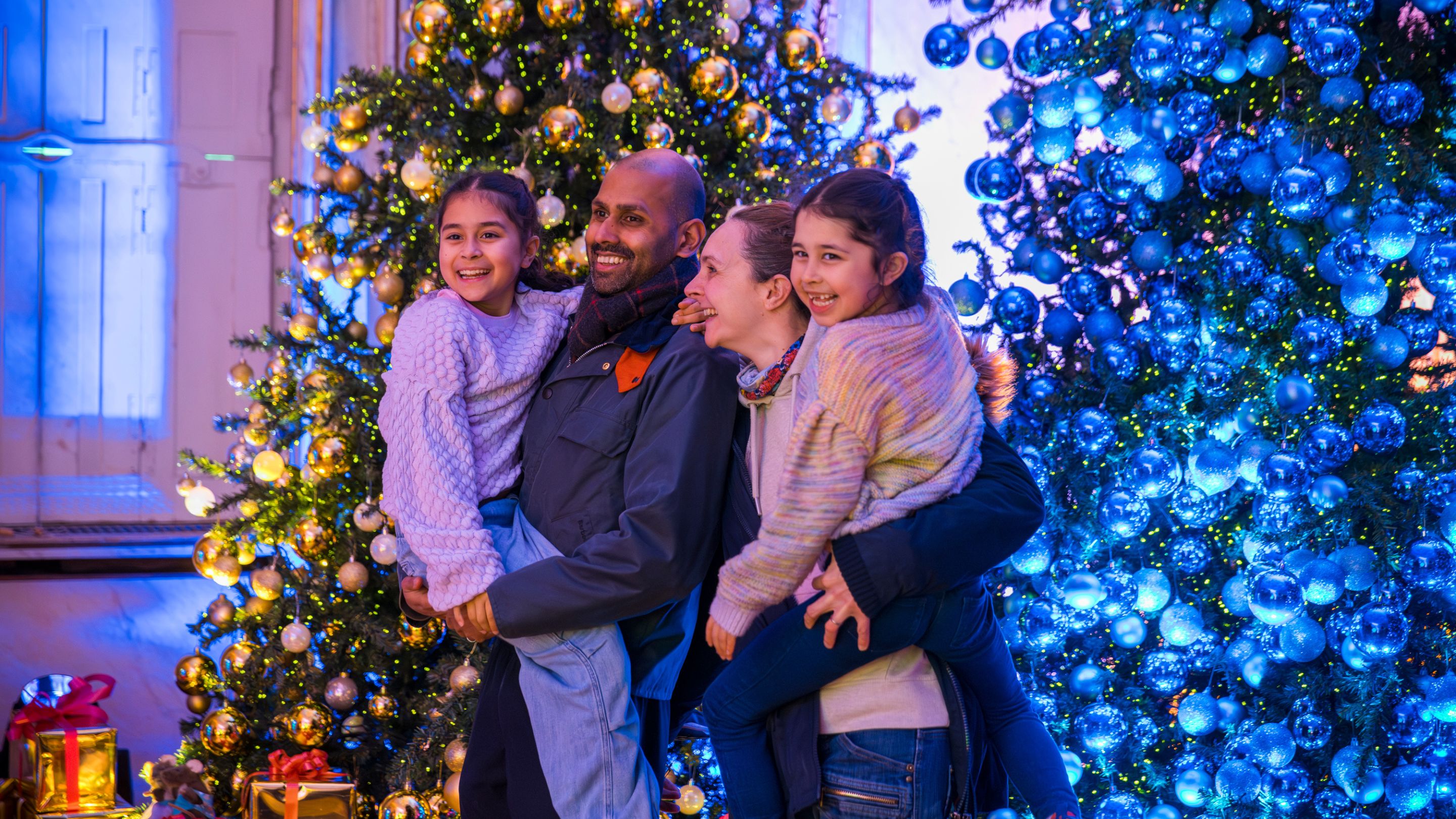 Man and woman both holding a young girl, standing smiling into the distance in Mottisfont's dining room, with decorated Christmas trees in backgroune