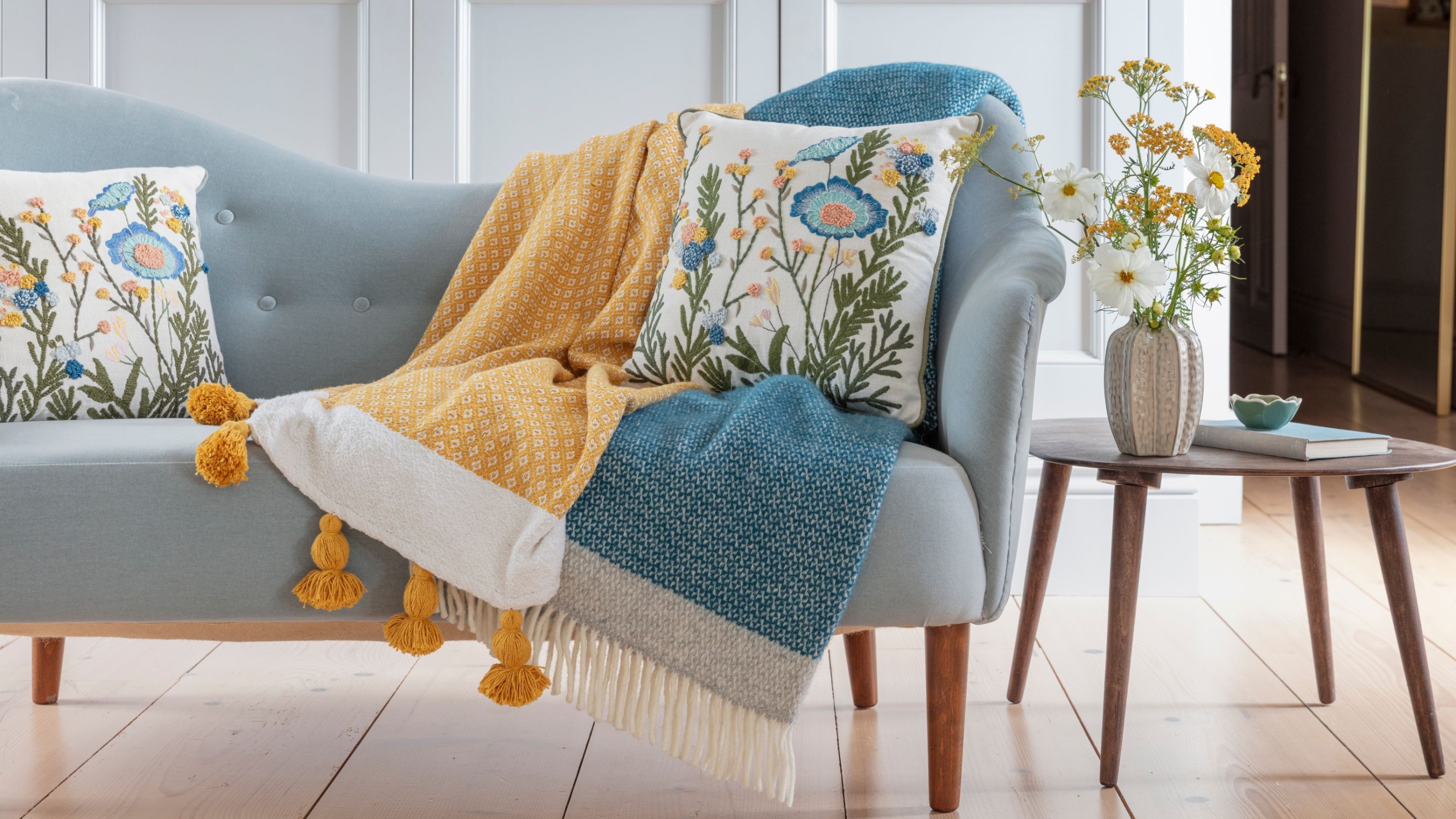 Assortment of spring 2026 retail items in pale blues and yellows including blankets and cushions displayed on a blue sofa in a room