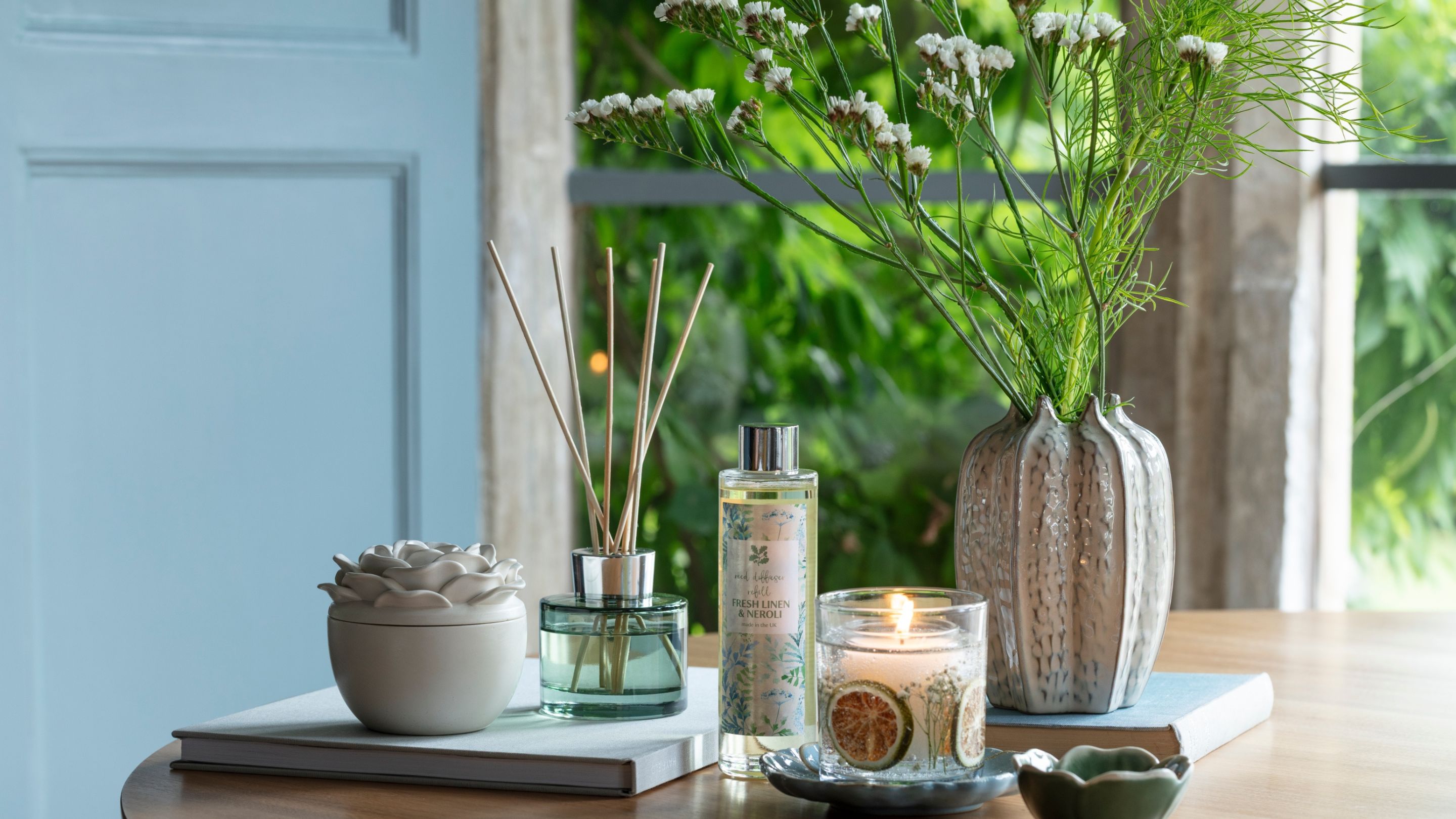 A room diffuser, candle and spray, together with some ceramic items and a vase of white flowers on a table with pale blue woodwork in background