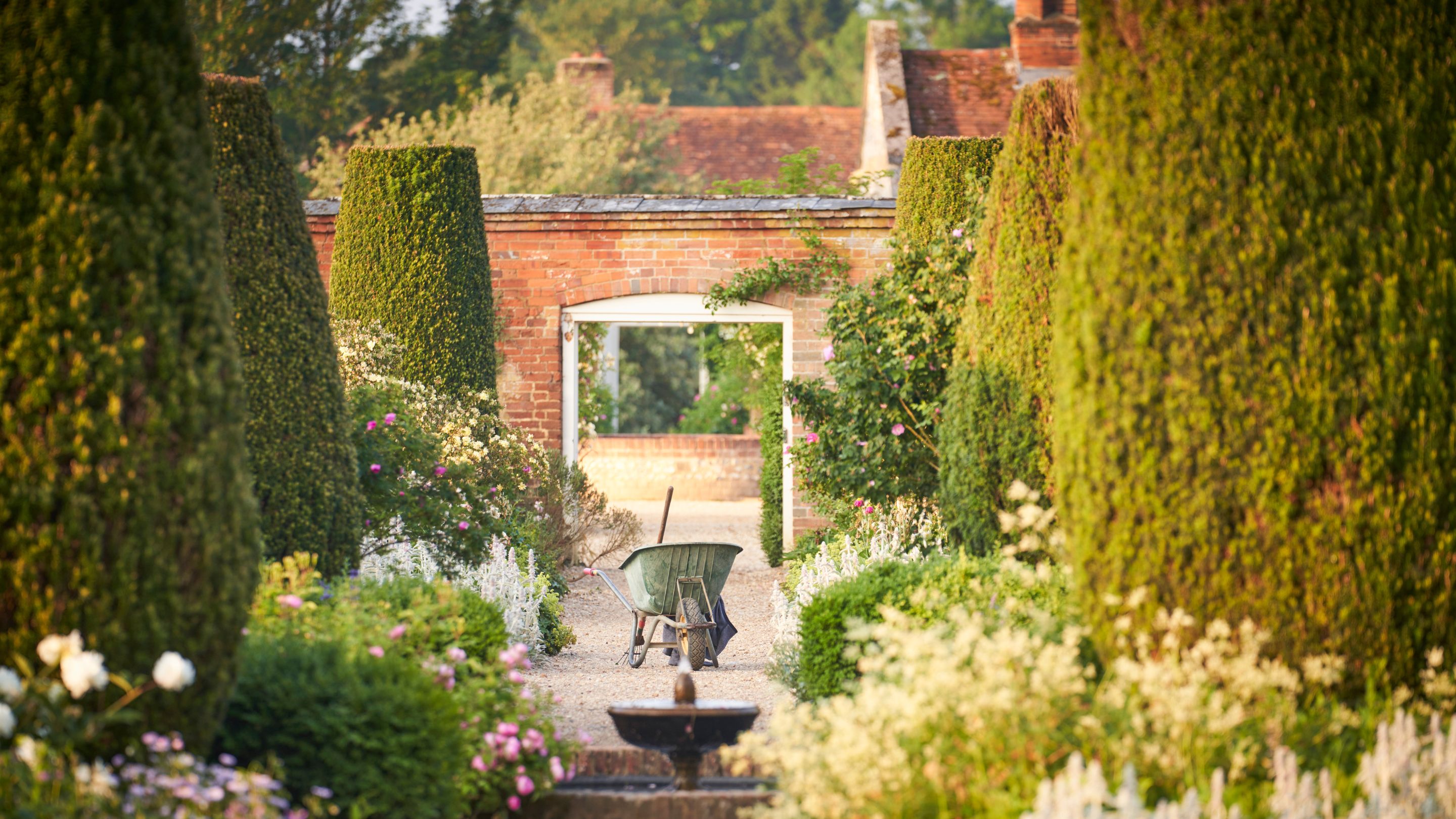 Gravel path with big flower borders on either side, within a walled garden, with mini water feature and doorway in wall in the background
