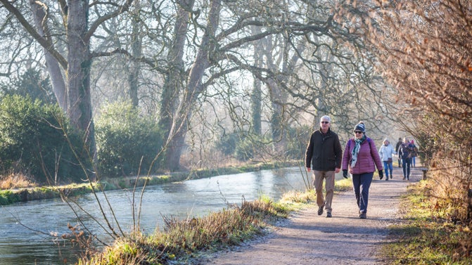 Two visitors walk towards the camera on a path leading alongside the straight banks of the River Test at Mottisfont. The visitors are wrapped up warm in coats for winter and other visitors can be seen following the same path behind them.