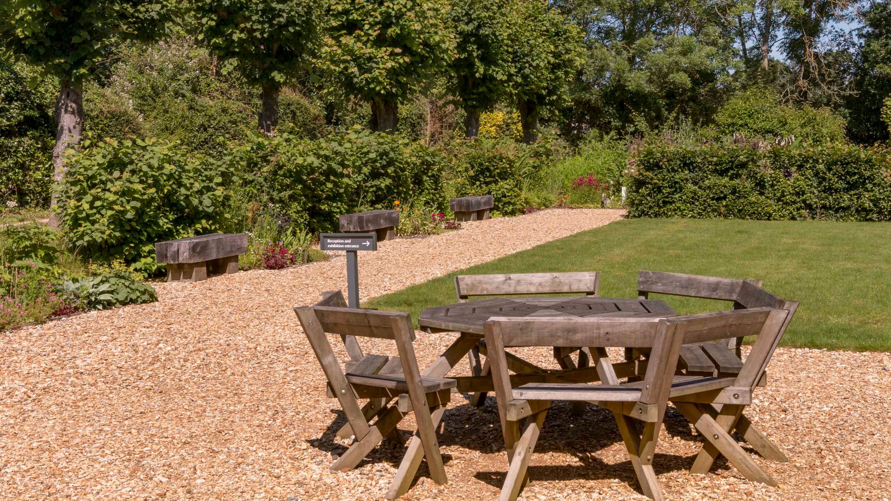 A wooden table and chairs on a gravel path in the rear gardens in summer at Sandham Memorial Chapel, Hampshire