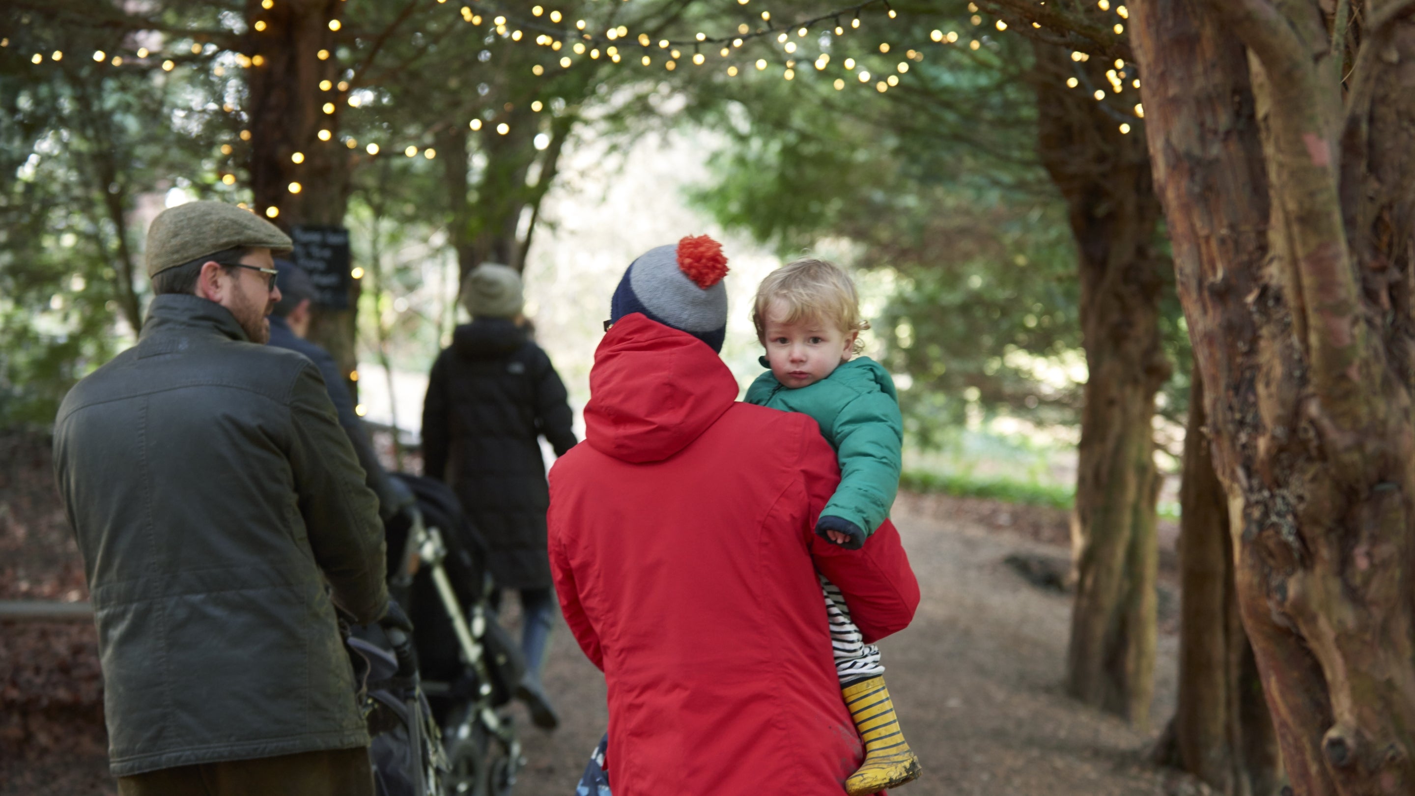 A man pushes a buggy while a woman in a bright red coat carries a young child down a woodland path with fairy lights overhead.