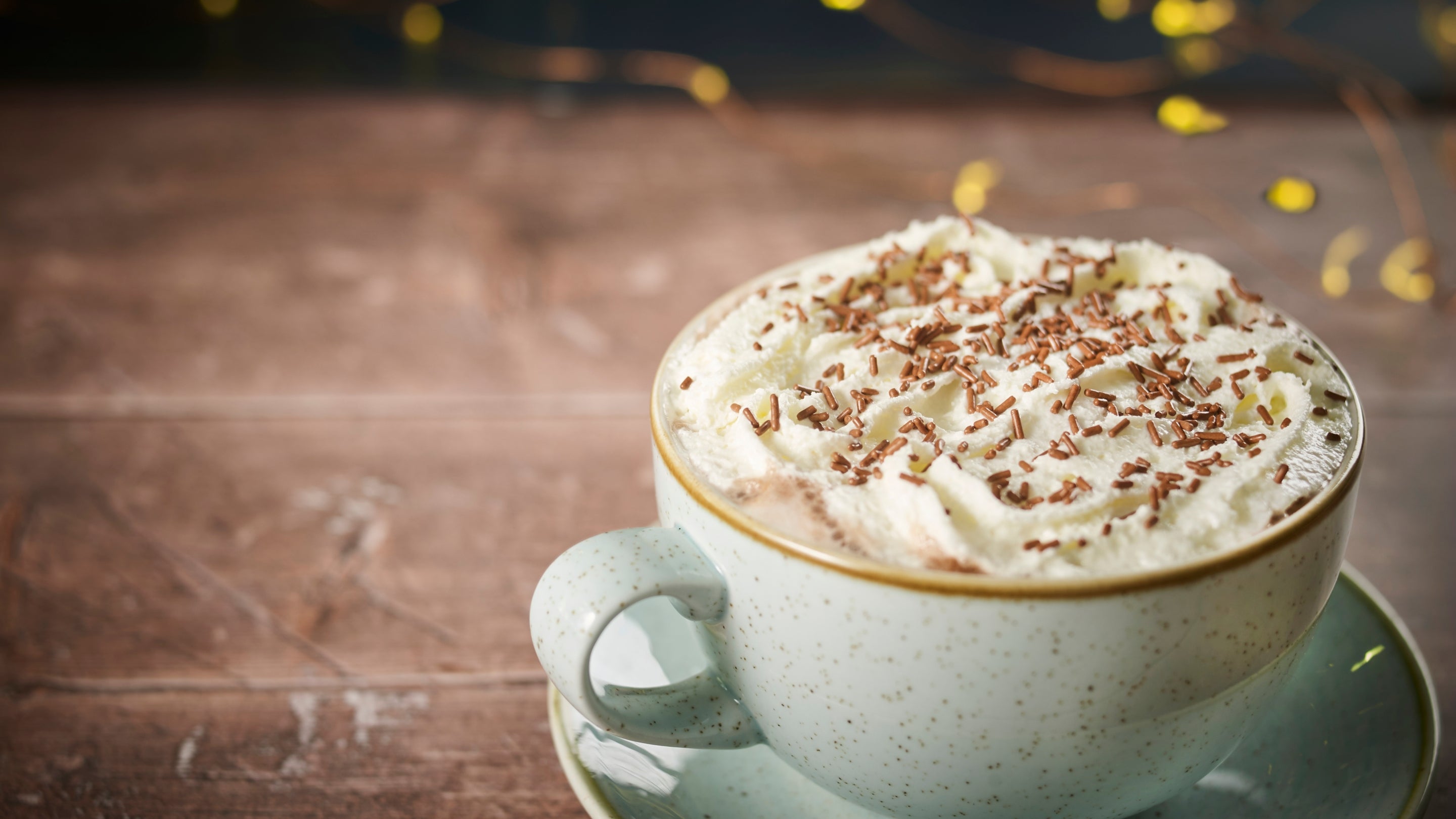 A cup of coconut hot chocolate with a topping of whipped cream is positioned bottom-right on a wooden surface with yellow lights out of focus in the background.