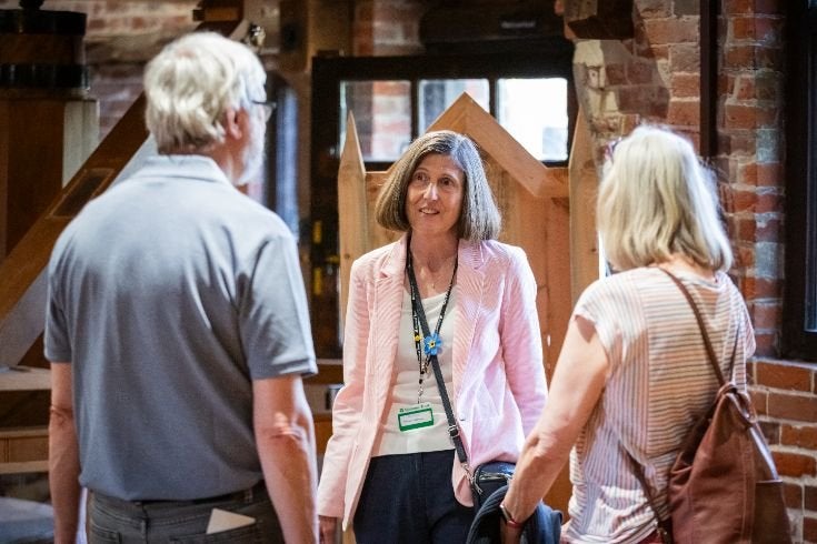 A person wearing a light pink jacket and a lanyard stands indoors in front of a wooden structure, speaking with two other people who are facing toward the camera. The setting appears to be a brick-walled interior space with warm lighting and visible wooden architectural elements.