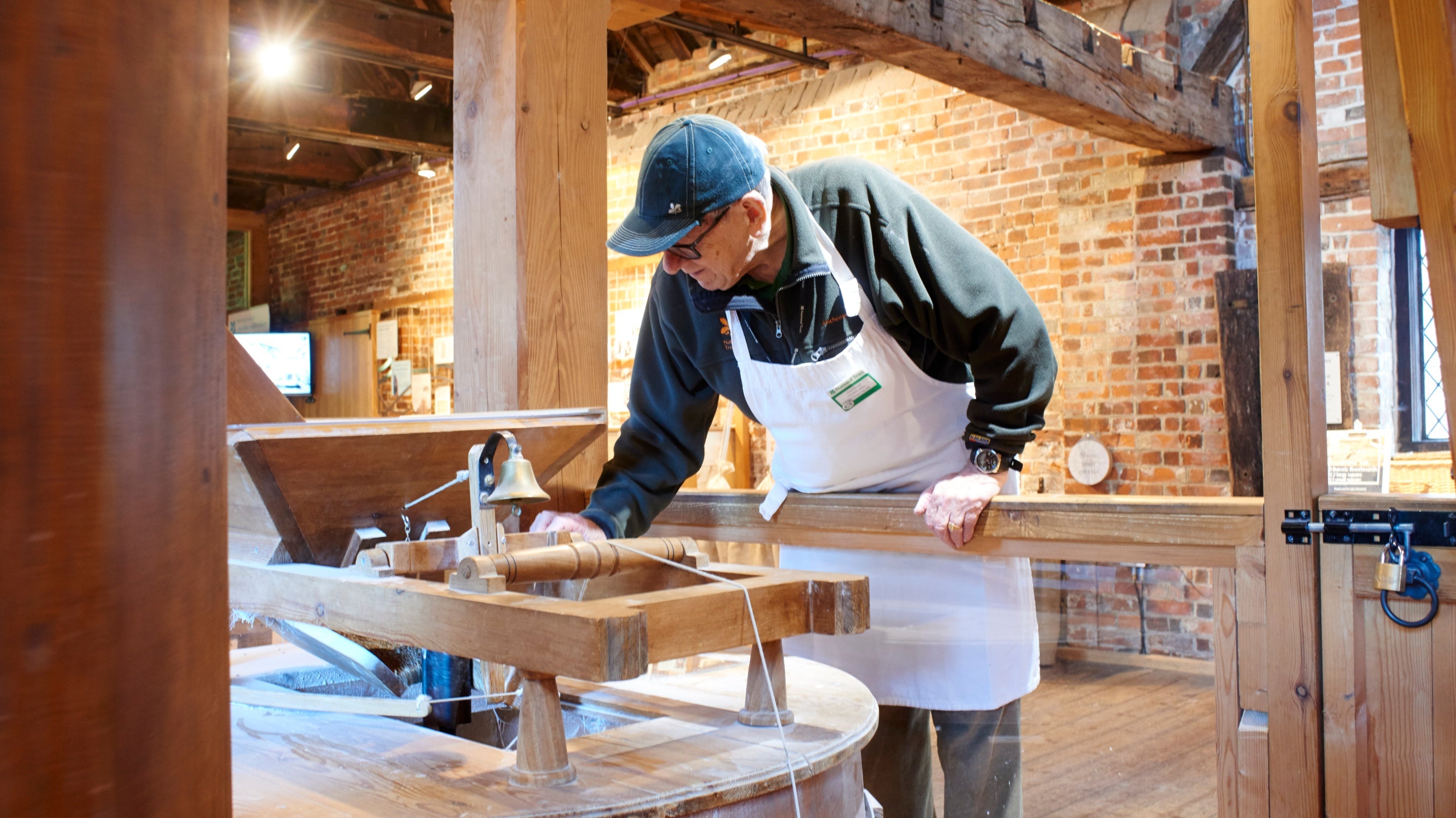 Volunteer wearing a cap and apron works with wooden machinery, overseeing the flour-making processes at Winchester City Mill, Hampshire