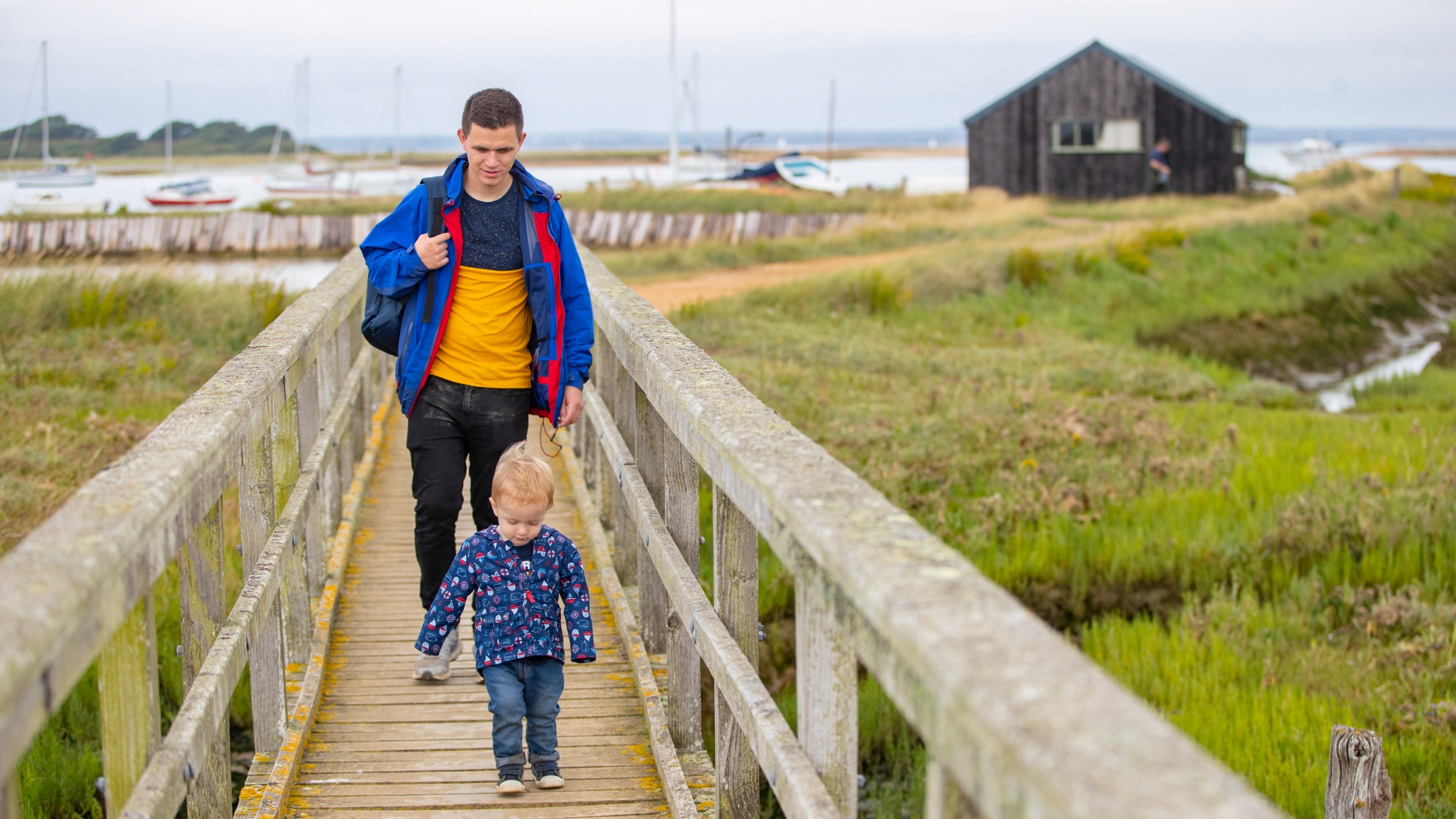 Family visitors walking along the boardwalk with a black boat house behind at Newtown National Nature Reserve.