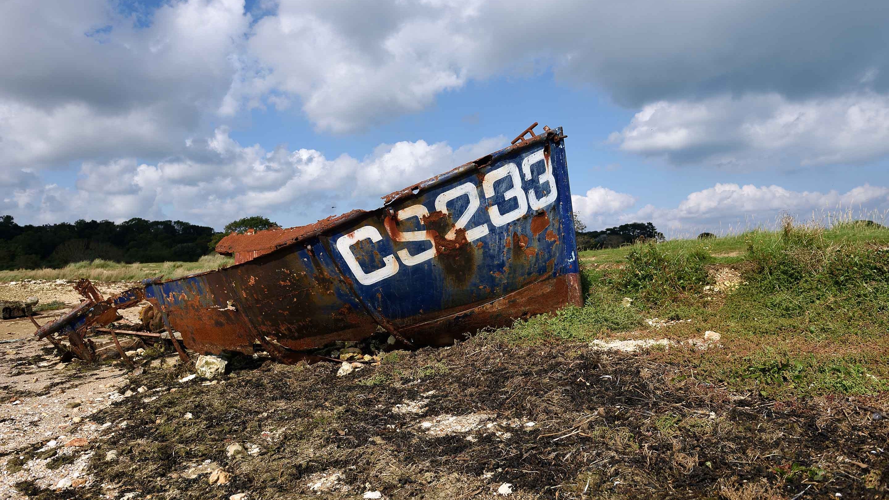 A rusted boat with 'CS233' painted on it in white lettering lies wrecked on a patch of sand and grass at St Helens Duver, Isle of Wight.