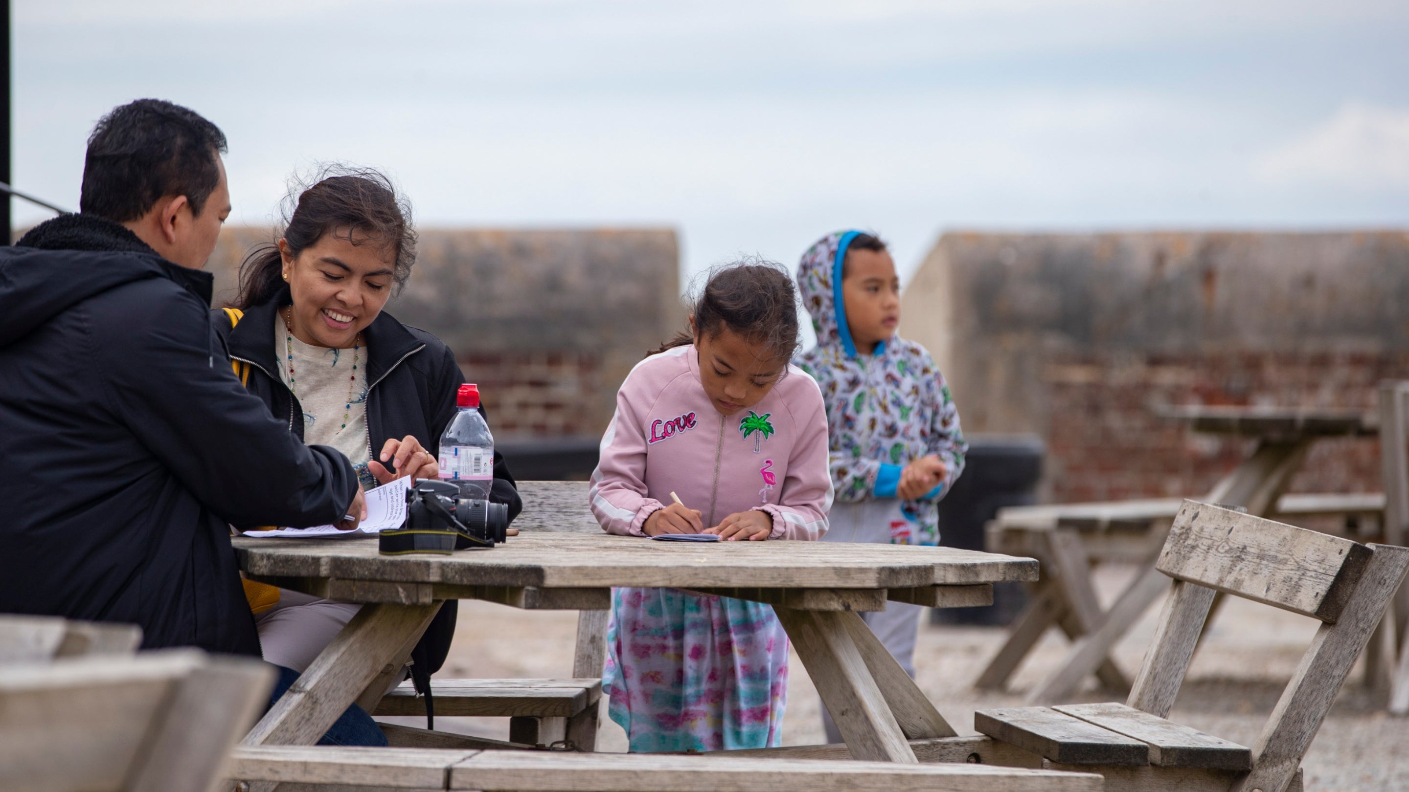 A family wearing coats sit at a wooden picnic table, with stone battlements behind them