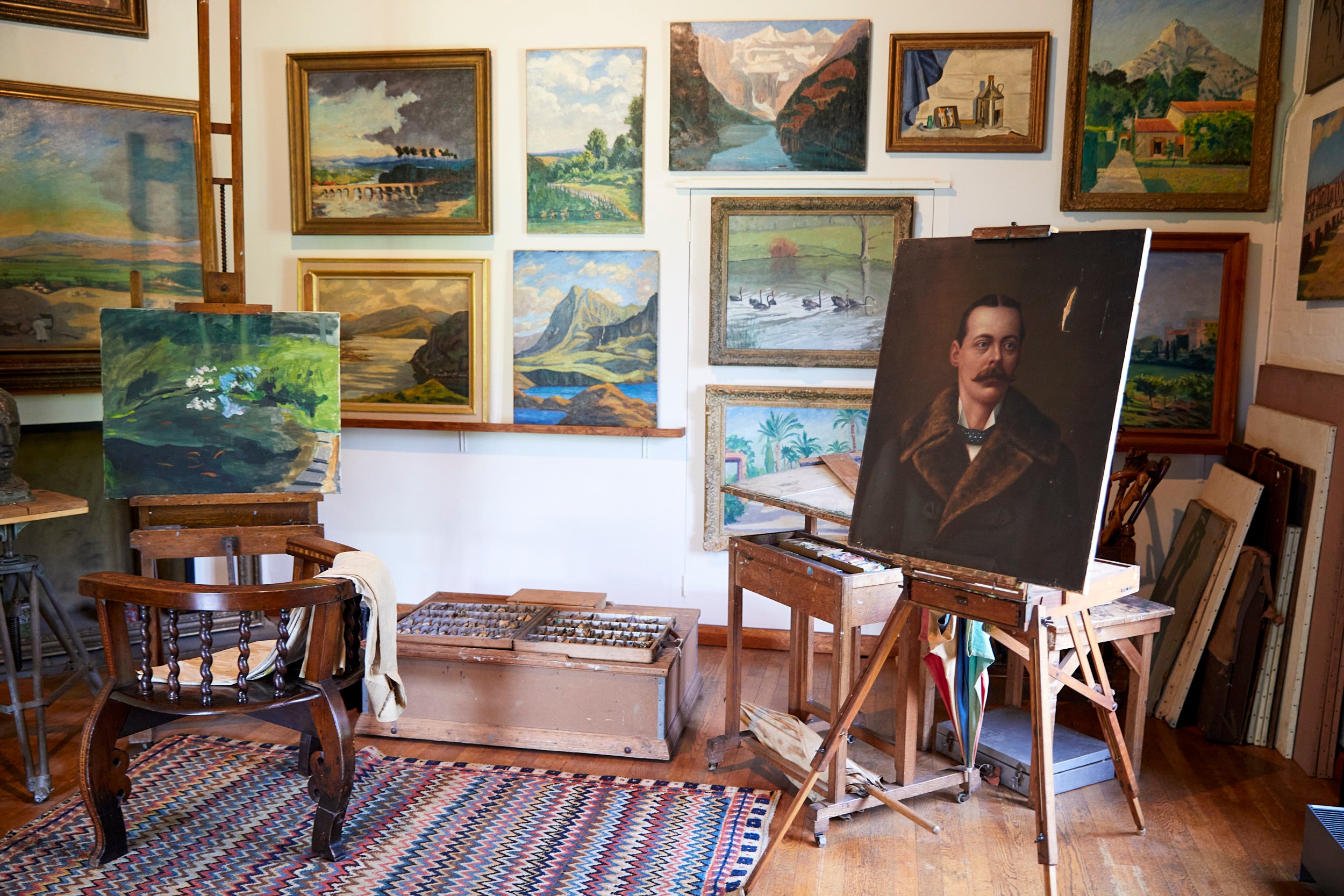 Interior of Winston S. Churchill's studio at Chartwell, Kent. Paintings by Churchill displayed on walls. Box of paints on the floor. Chair draped with a beige jacket sits in front of  and easel with painting of the golden orfe pond. Another portrait stands on an easel.