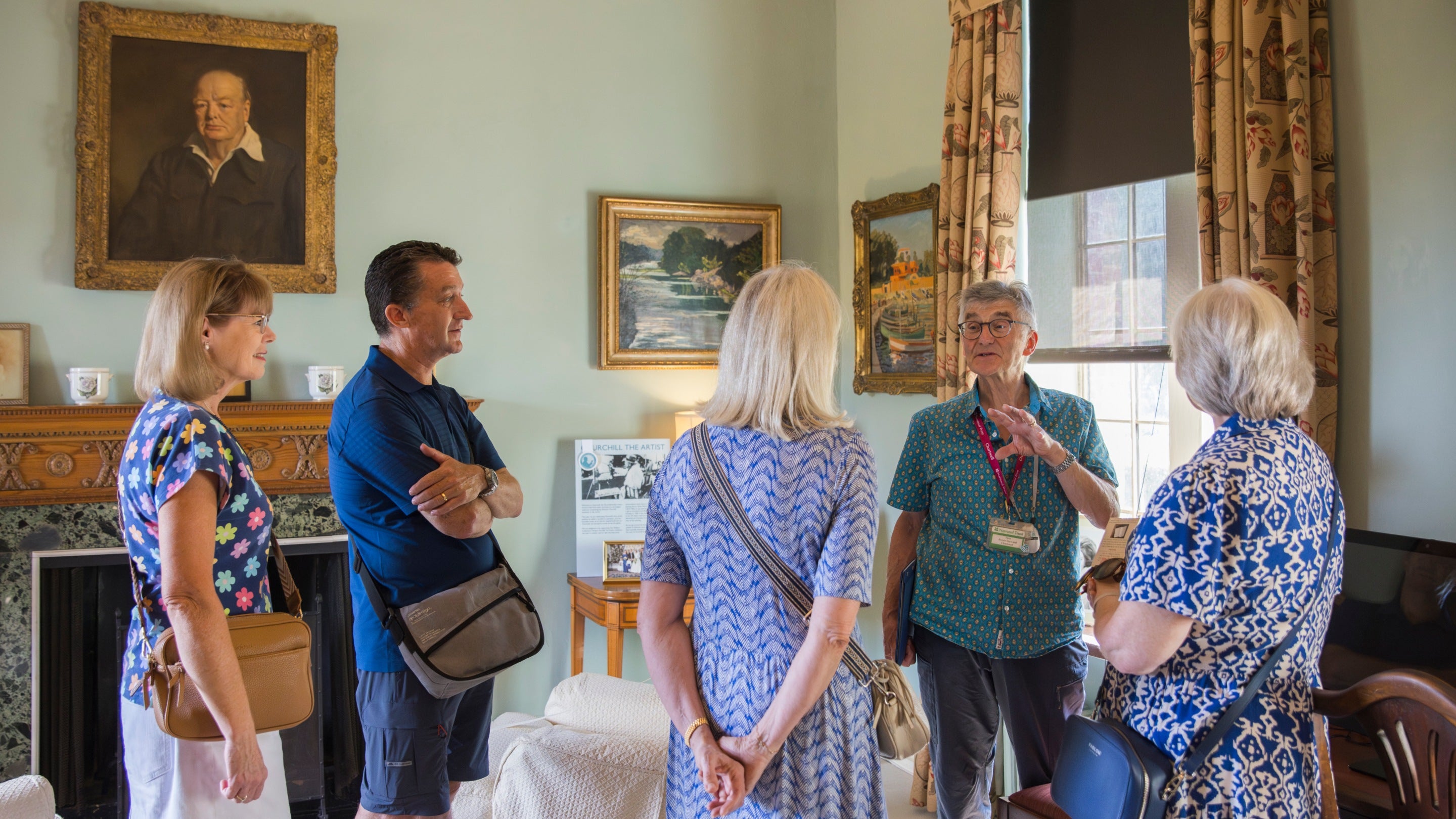 4visitors, 3 women and 1 man listening to a Chartwell room steward. They are in the Sitting Room. A portrait of Churchill hangs above the mantlepiece in the background.